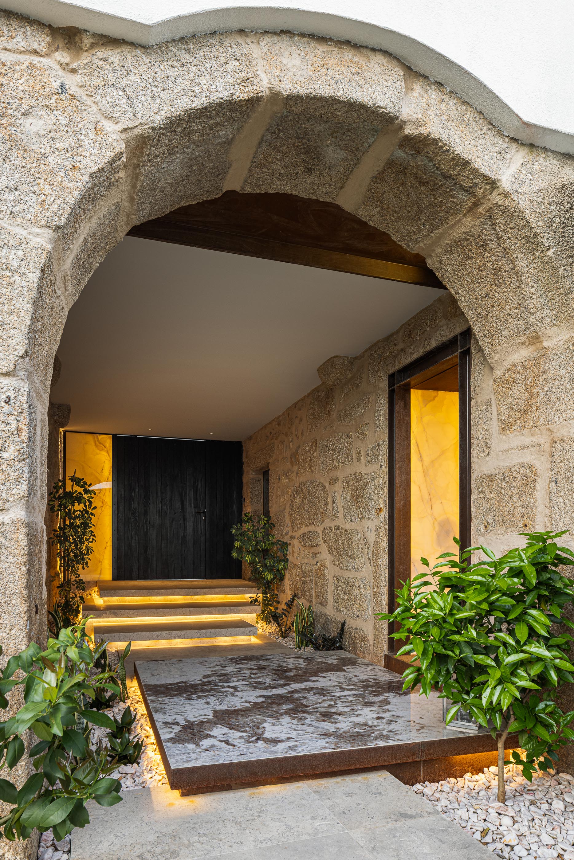 Entrada principal: Arco de pedra rústica, porta escura, iluminação embutida e vegetação exuberante. — Moradia do Retiro arquitetura Ricardo Azevedo Arquitecto em Santo Tirso