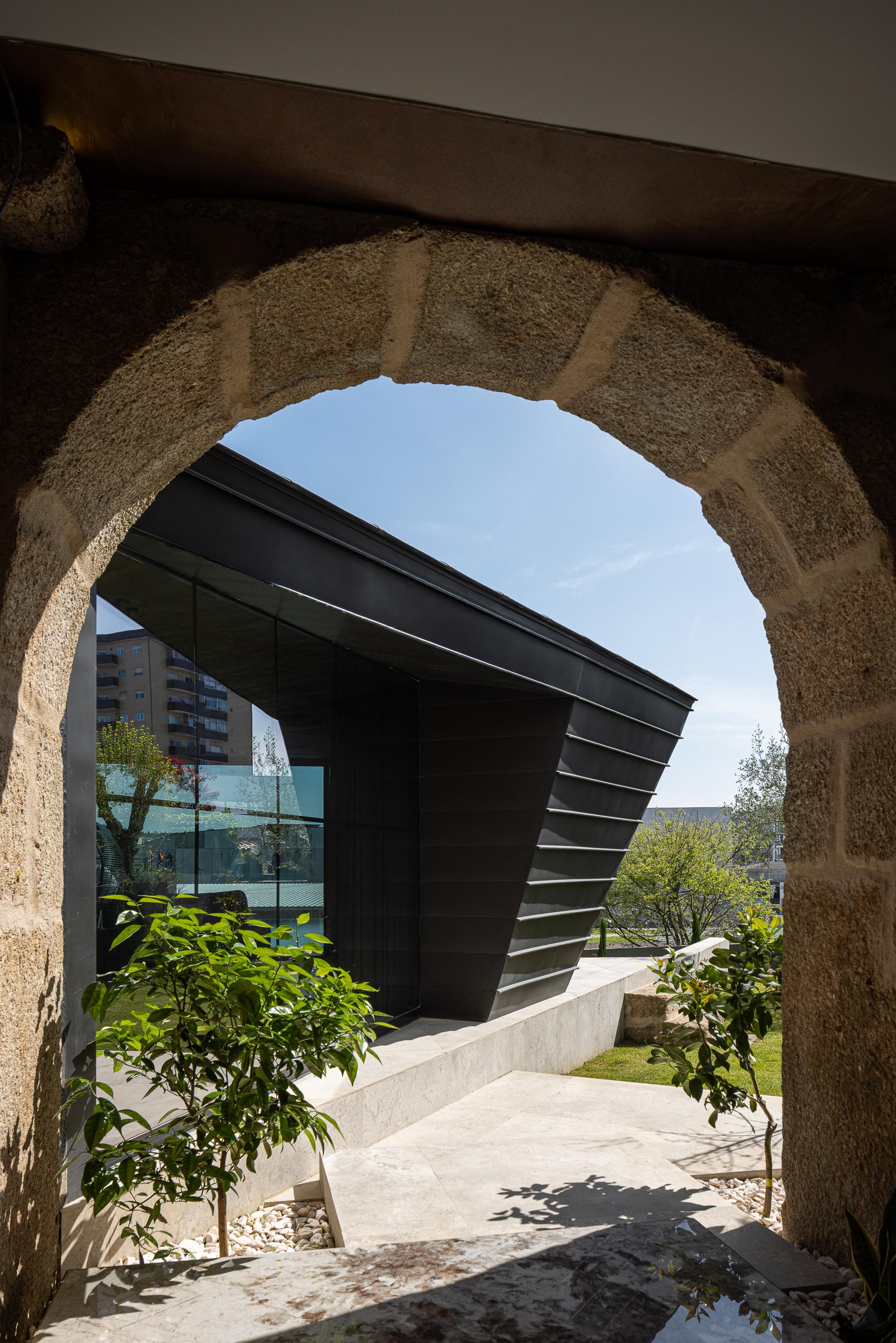 Exterior: Vista através de arco de pedra para o anexo moderno com revestimento metálico preto e jardim. — Moradia do Retiro arquitetura Ricardo Azevedo Arquitecto em Santo Tirso