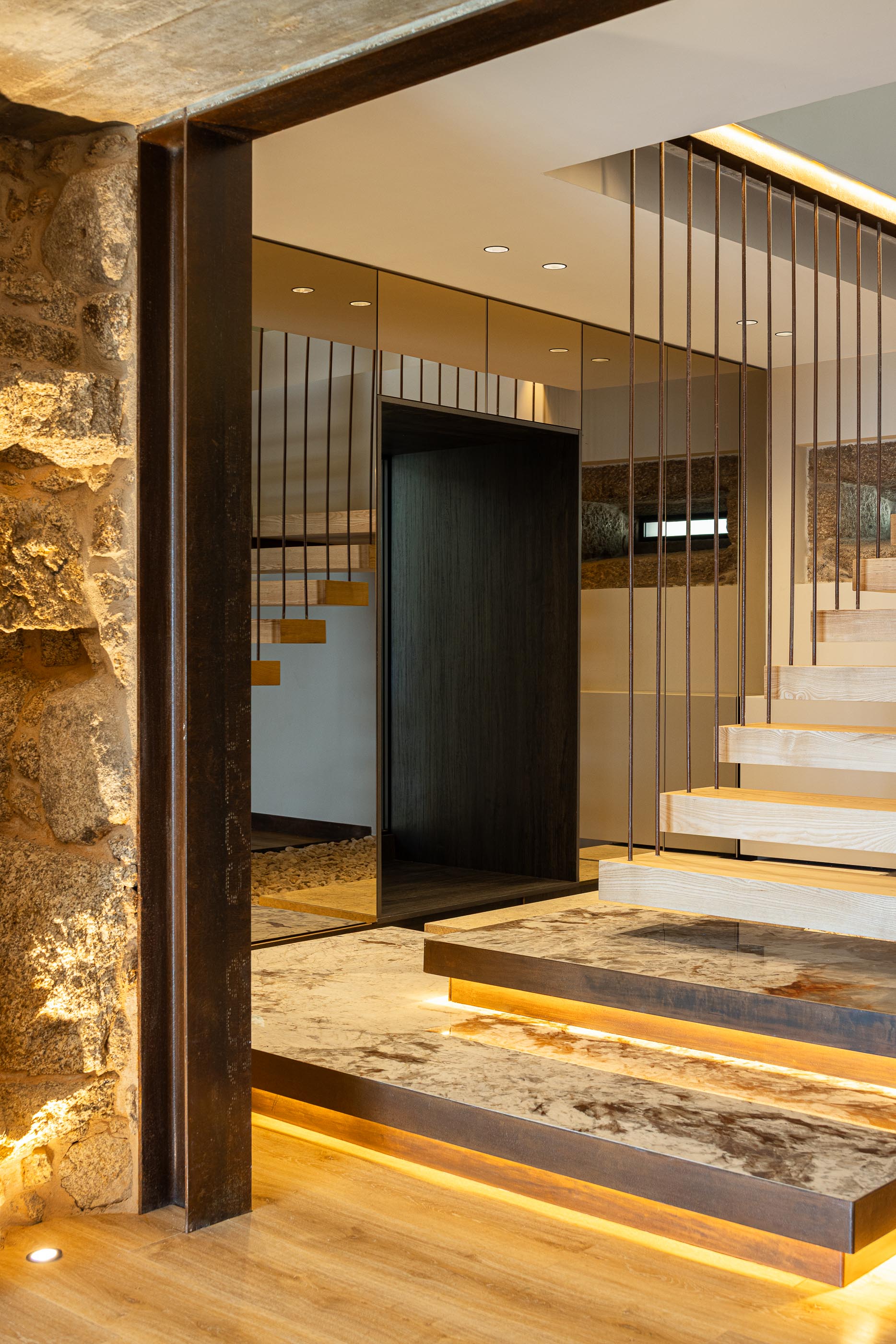 Escadas: Degraus iluminados, guarda em metal, parede em pedra rústica e espelho. — Moradia do Retiro arquitetura Ricardo Azevedo Arquitecto em Santo Tirso
