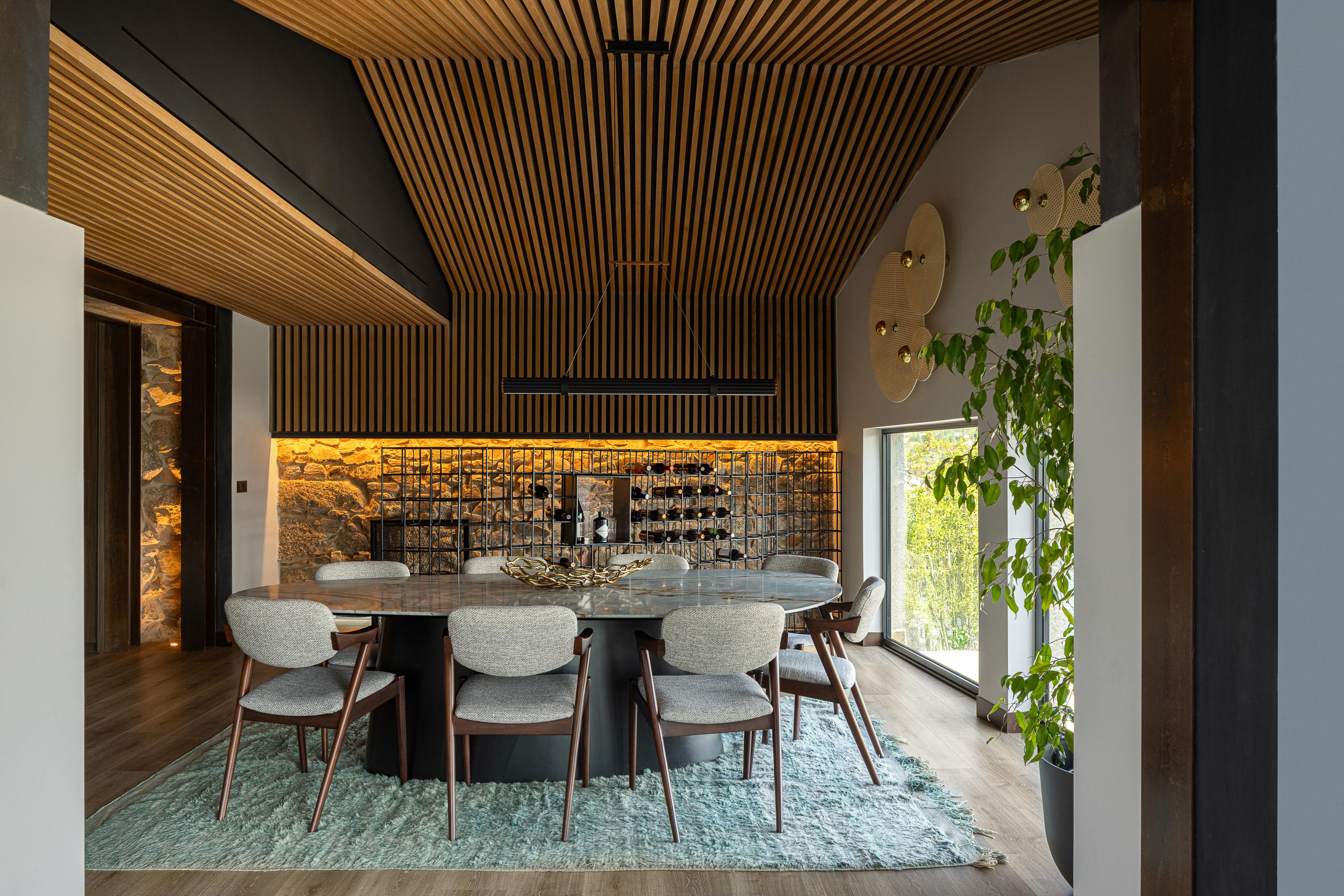 Sala de jantar: Mesa oval em pedra, cadeiras estofadas, garrafeira e teto em madeira com iluminação. — Moradia do Retiro arquitetura Ricardo Azevedo Arquitecto em Santo Tirso
