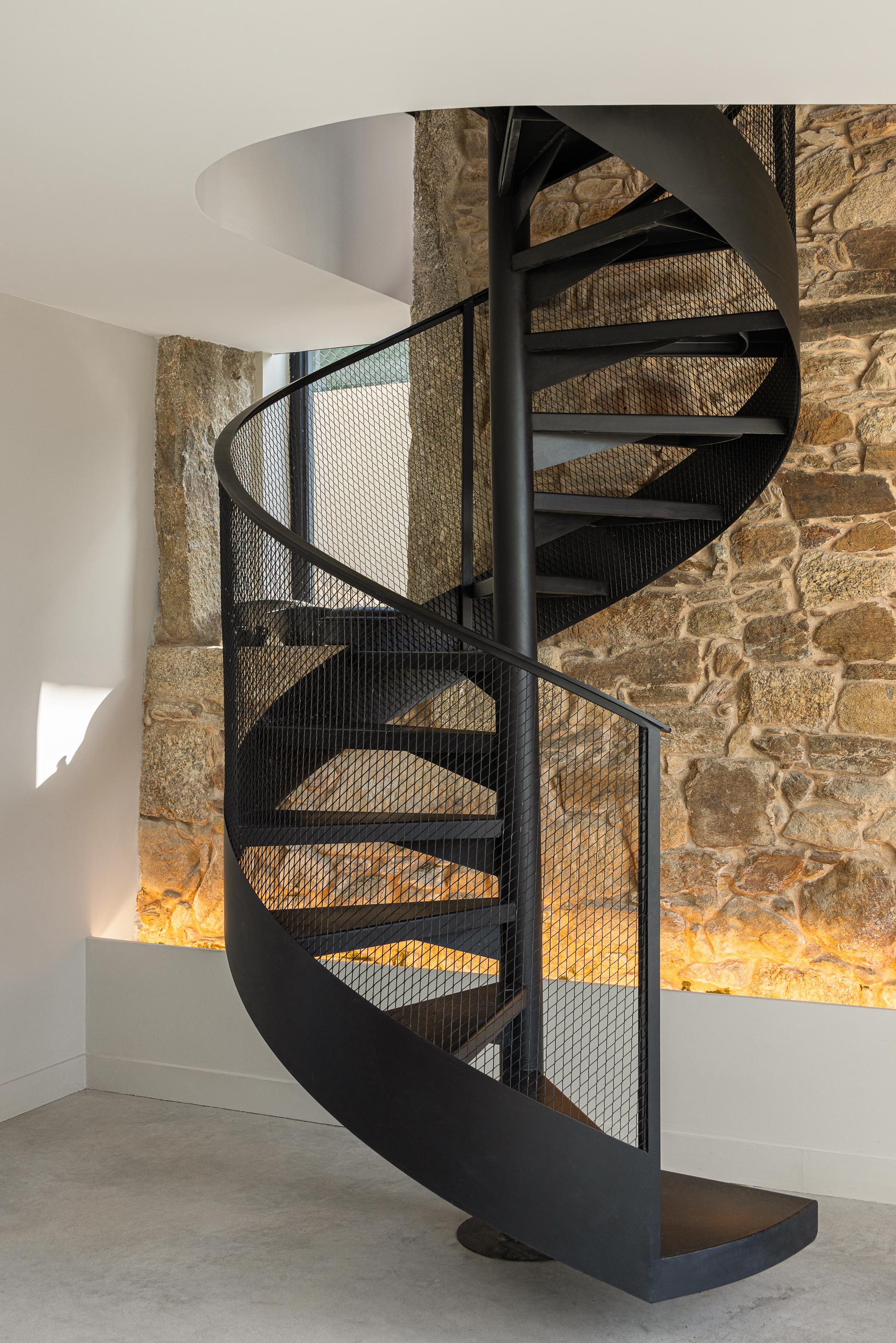 Escadas: Escada em caracol de metal preto junto a parede de pedra rústica com iluminação. — Moradia do Retiro arquitetura Ricardo Azevedo Arquitecto em Santo Tirso