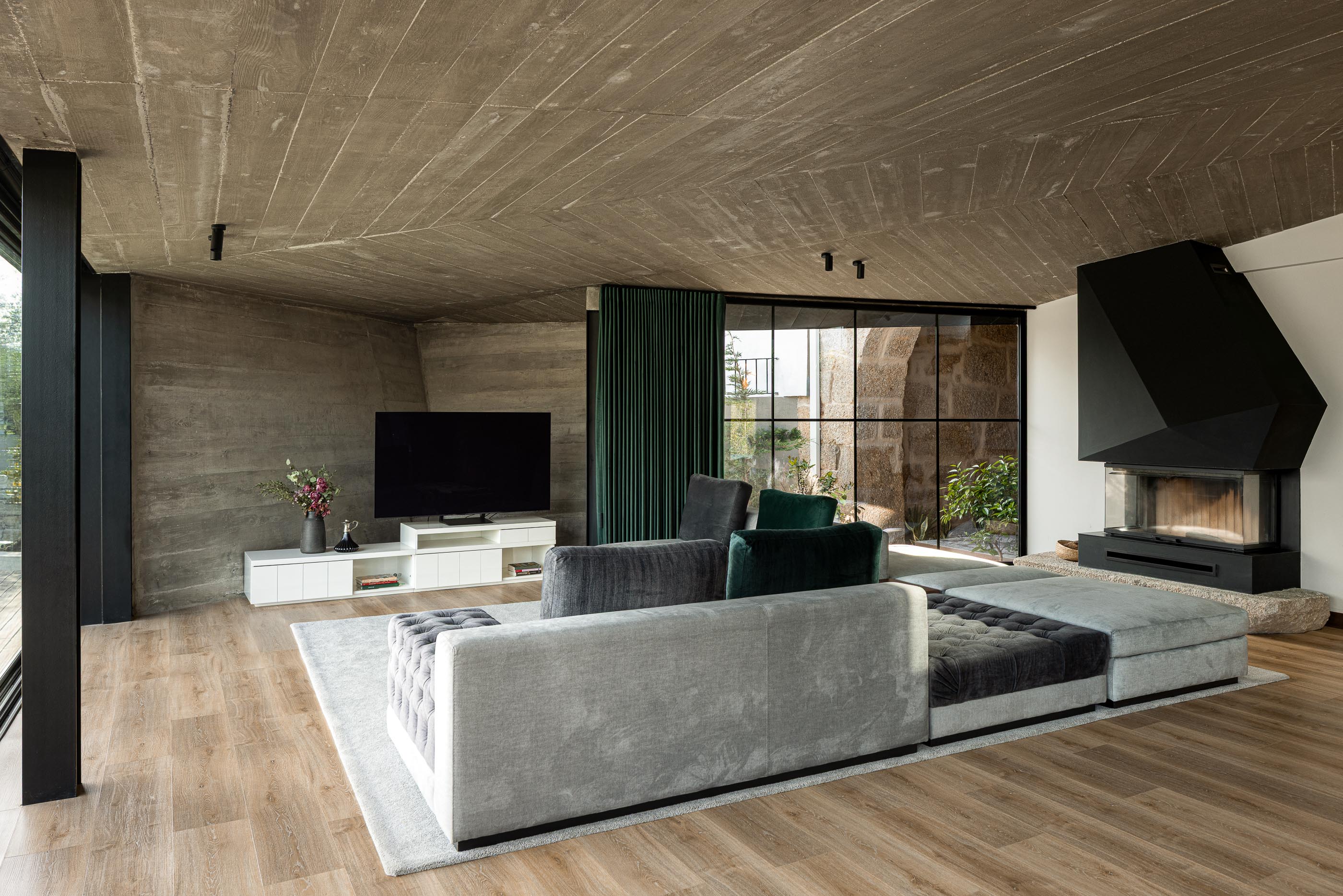 Sala de estar: Sofá cinza, lareira preta geométrica, teto em betão aparente e parede em terra batida. — Moradia do Retiro arquitetura Ricardo Azevedo Arquitecto em Santo Tirso
