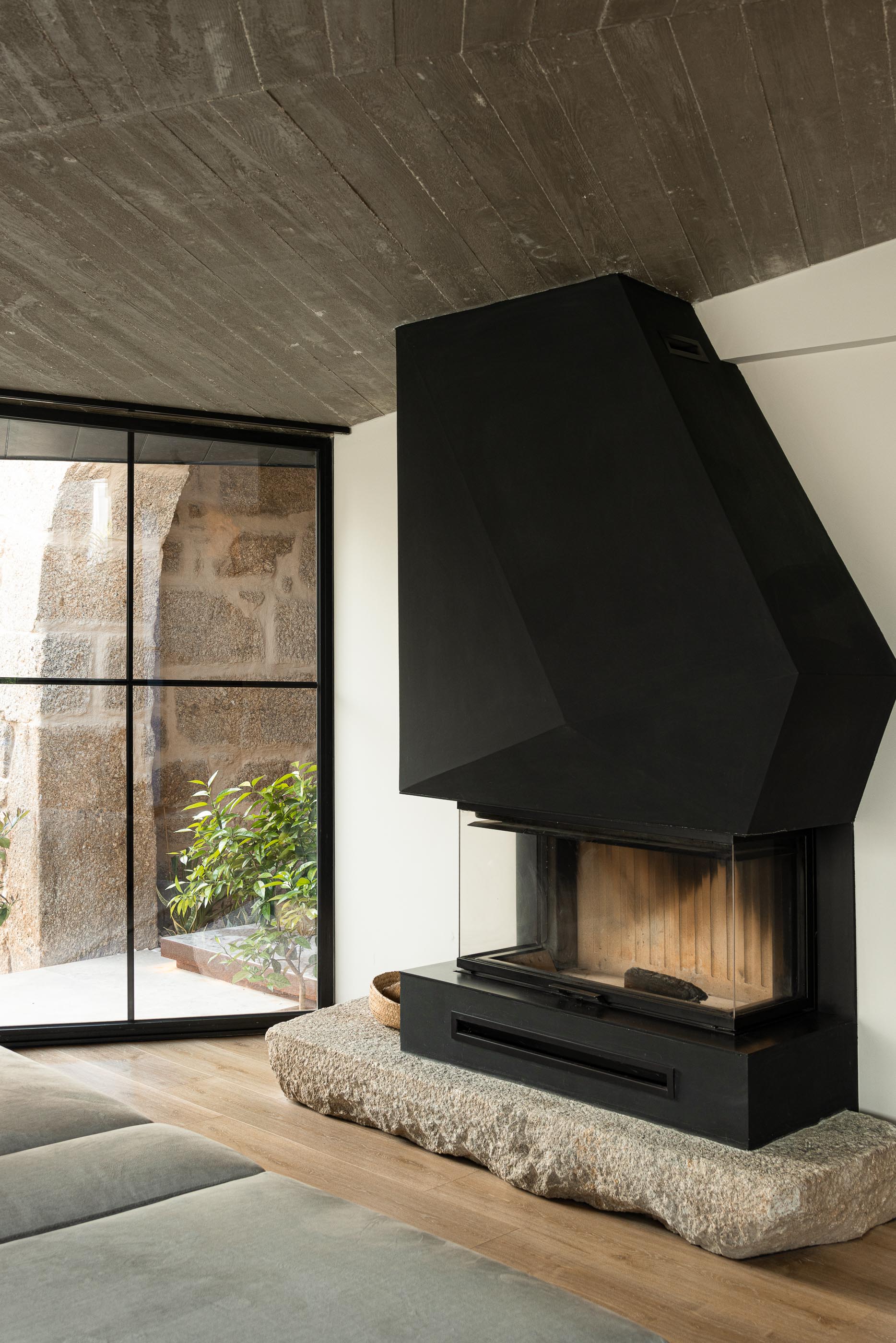 Sala de estar: Lareira preta geométrica sobre base de pedra, janela com vista para o jardim interior. — Moradia do Retiro arquitetura Ricardo Azevedo Arquitecto em Santo Tirso