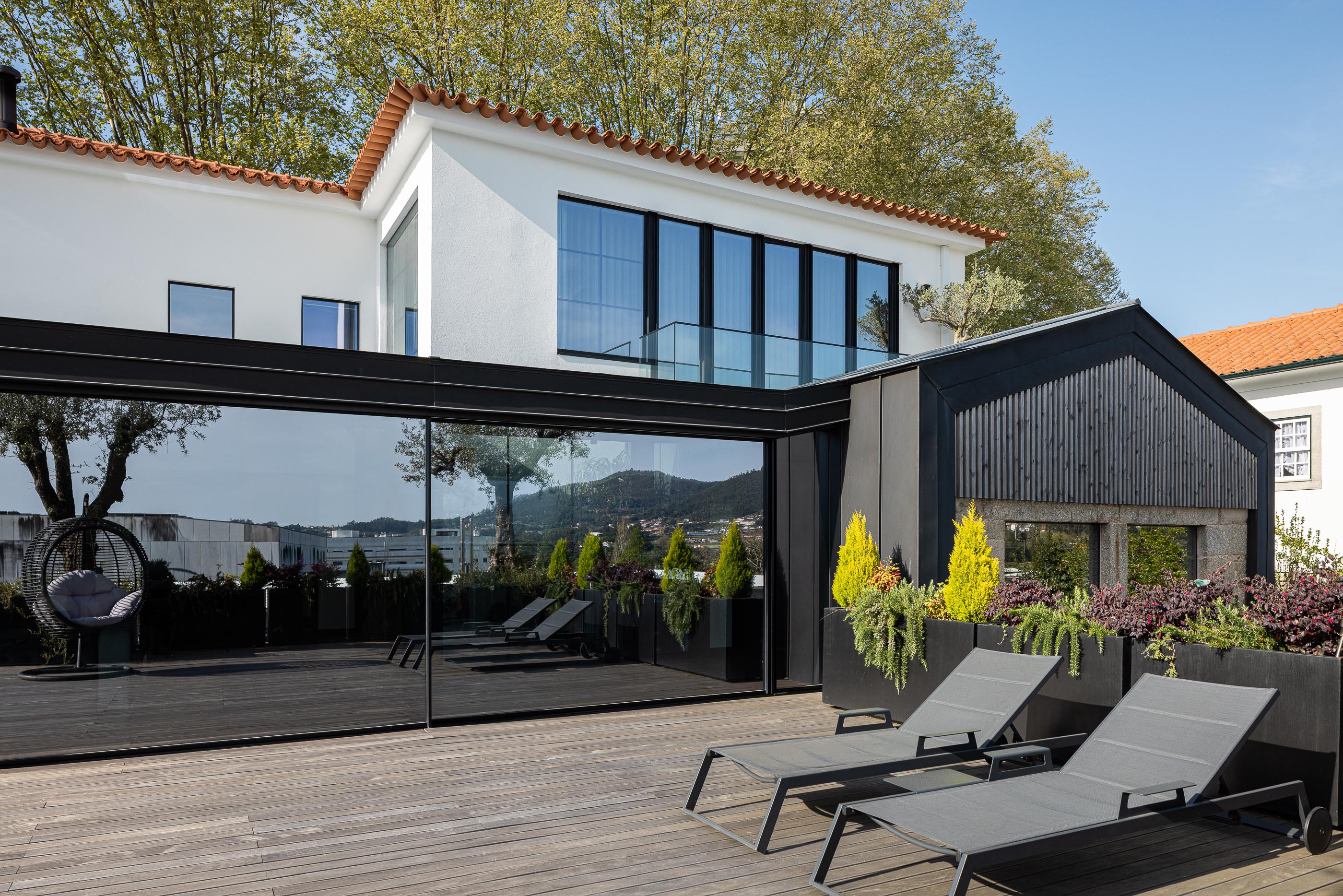 Terraço: Espreguiçadeiras, plantas em vasos, anexo moderno e fachada da moradia. — Moradia do Retiro arquitetura Ricardo Azevedo Arquitecto em Santo Tirso