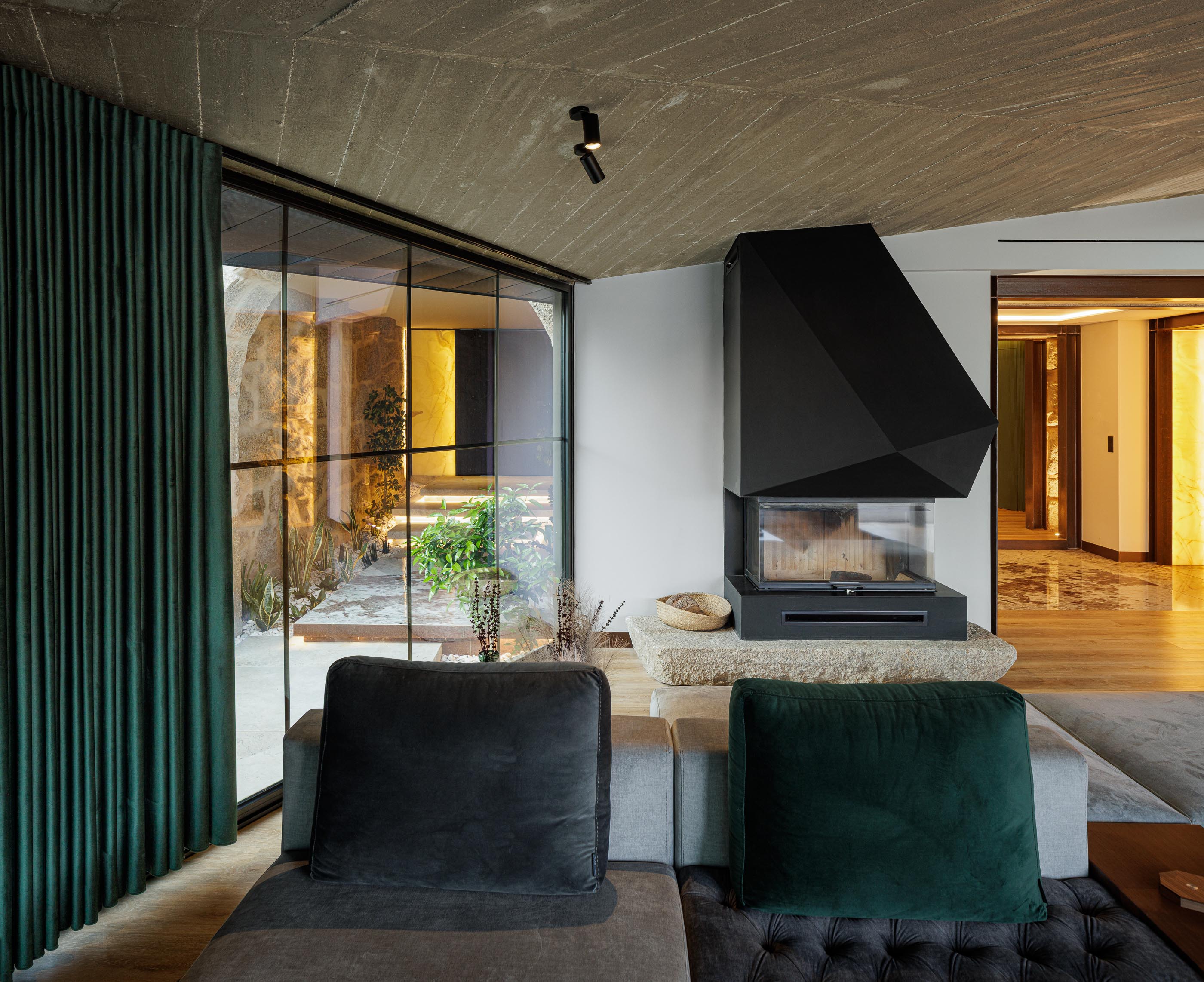Sala de estar: Lareira preta, sofá cinza, teto em betão aparente e vista para o jardim interior. — Moradia do Retiro arquitetura Ricardo Azevedo Arquitecto em Santo Tirso