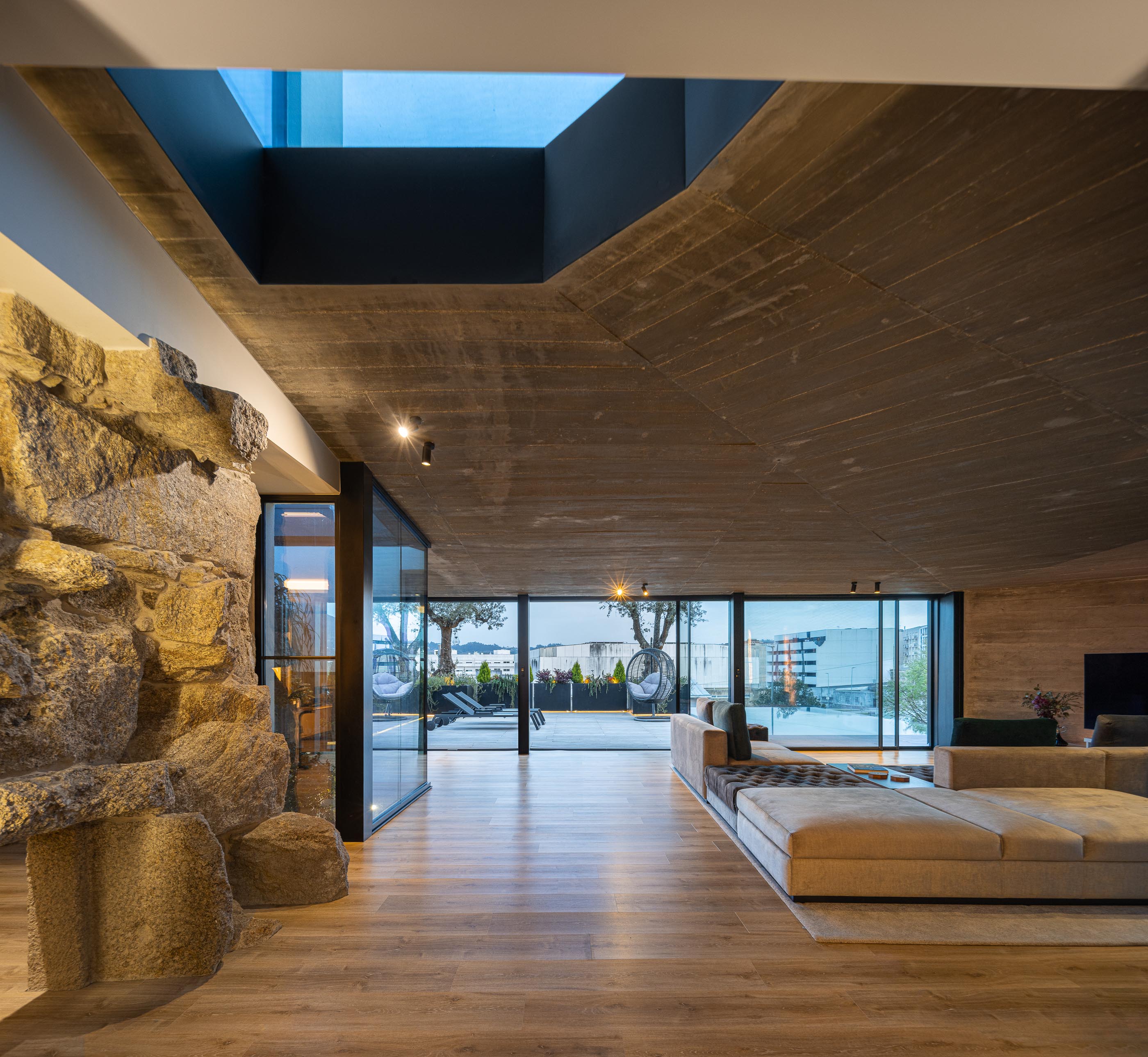 Sala de estar: Teto em betão aparente, parede em pedra rústica, grandes janelas e sofá modular. — Moradia do Retiro arquitetura Ricardo Azevedo Arquitecto em Santo Tirso