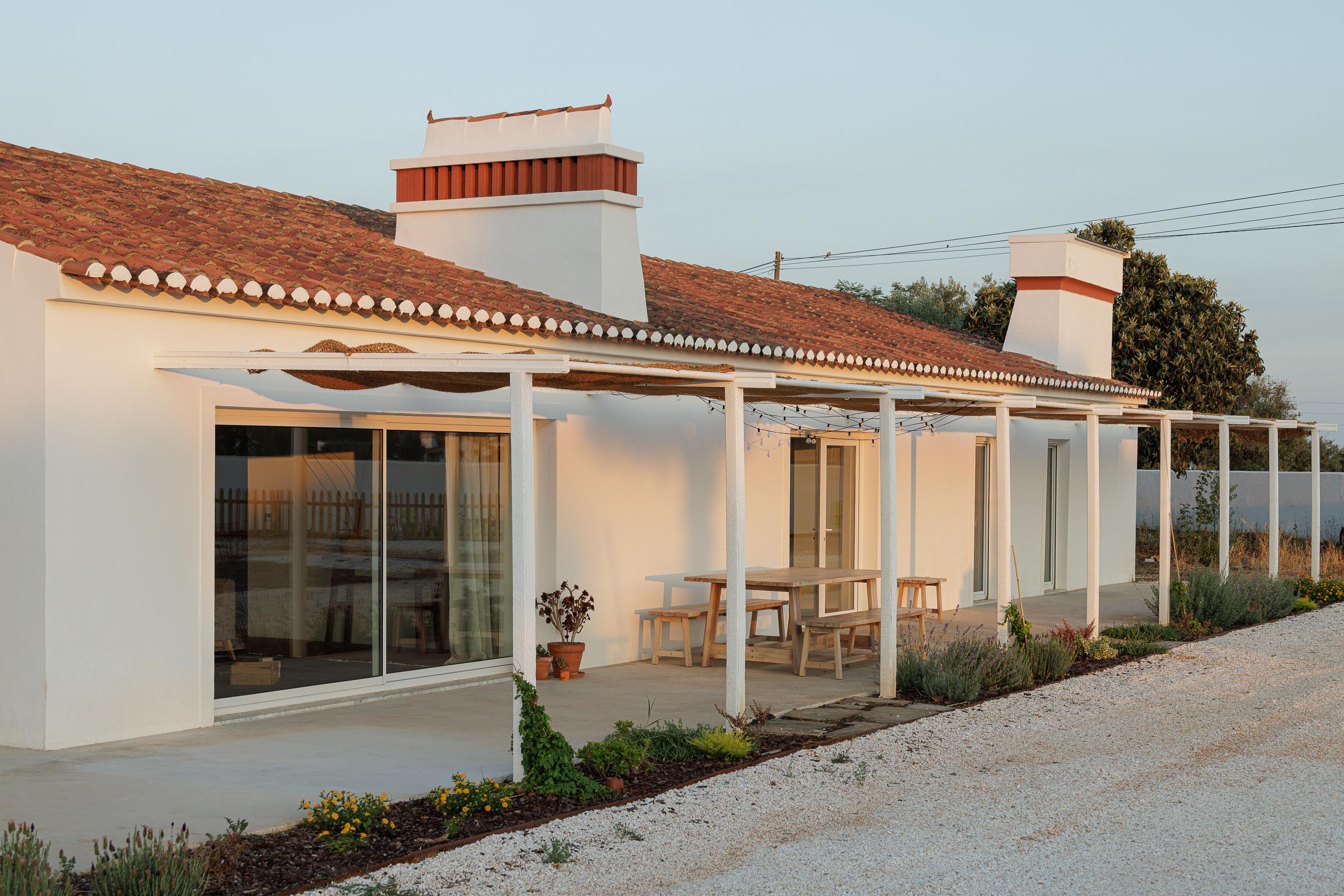 Exterior: Fachada branca com telhado de telha, alpendre com cobertura de palha, mesa de madeira e gravilha no pavimento. — Monte do Divor arquitetura Vasco Burnay Arquitectura em Estremoz