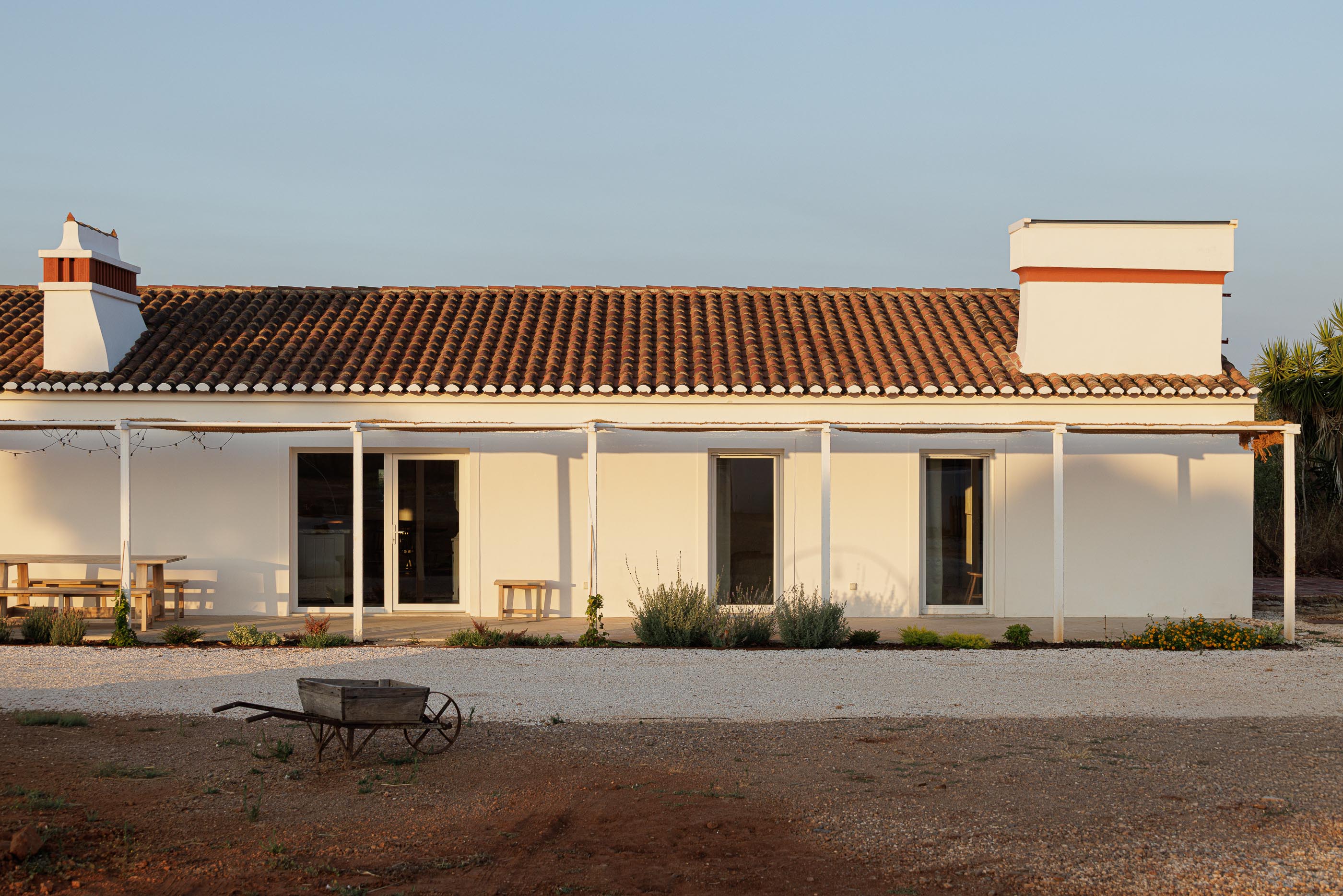 Exterior: Vista lateral da casa branca com telhado de telha, alpendre e terreno com gravilha. — Monte do Divor arquitetura Vasco Burnay Arquitectura em Estremoz