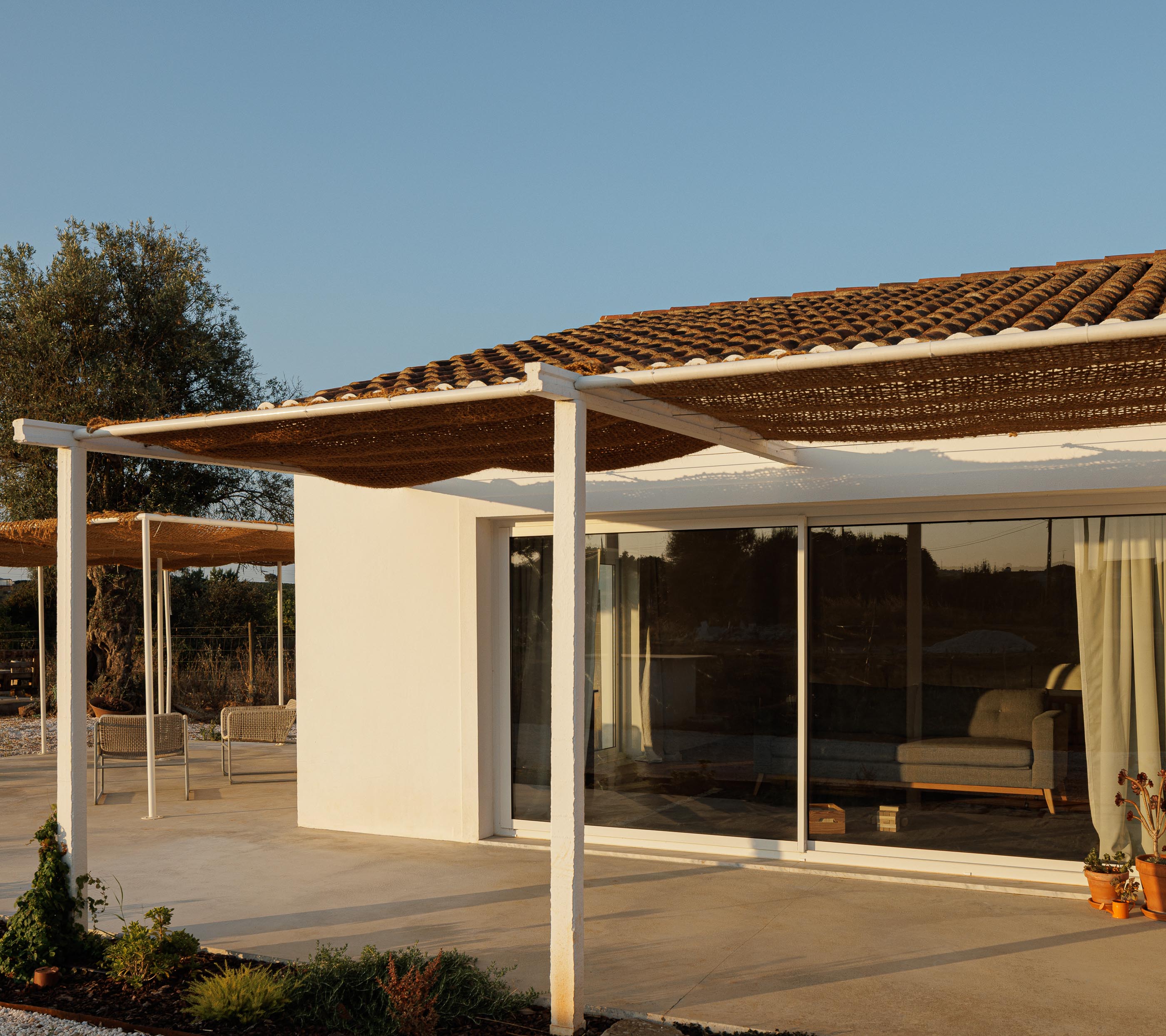 Exterior: Alpendre com pavimento em cimento, cadeiras de corda e vista para a sala de estar através da janela. — Monte do Divor arquitetura Vasco Burnay Arquitectura em Estremoz