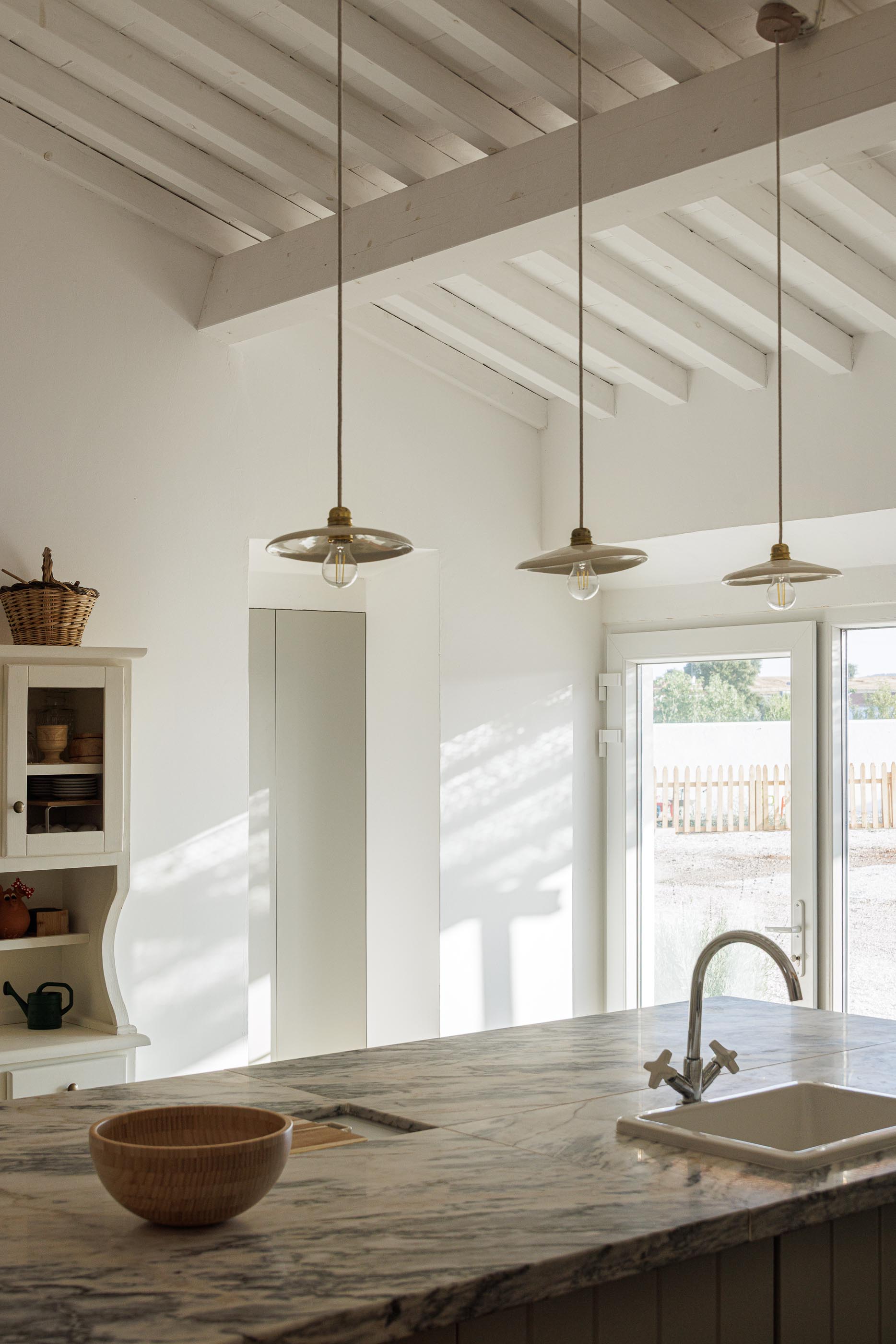 Cozinha: Ilha com bancada em mármore, candeeiros suspensos e teto branco com vigas de madeira. — Monte do Divor arquitetura Vasco Burnay Arquitectura em Estremoz