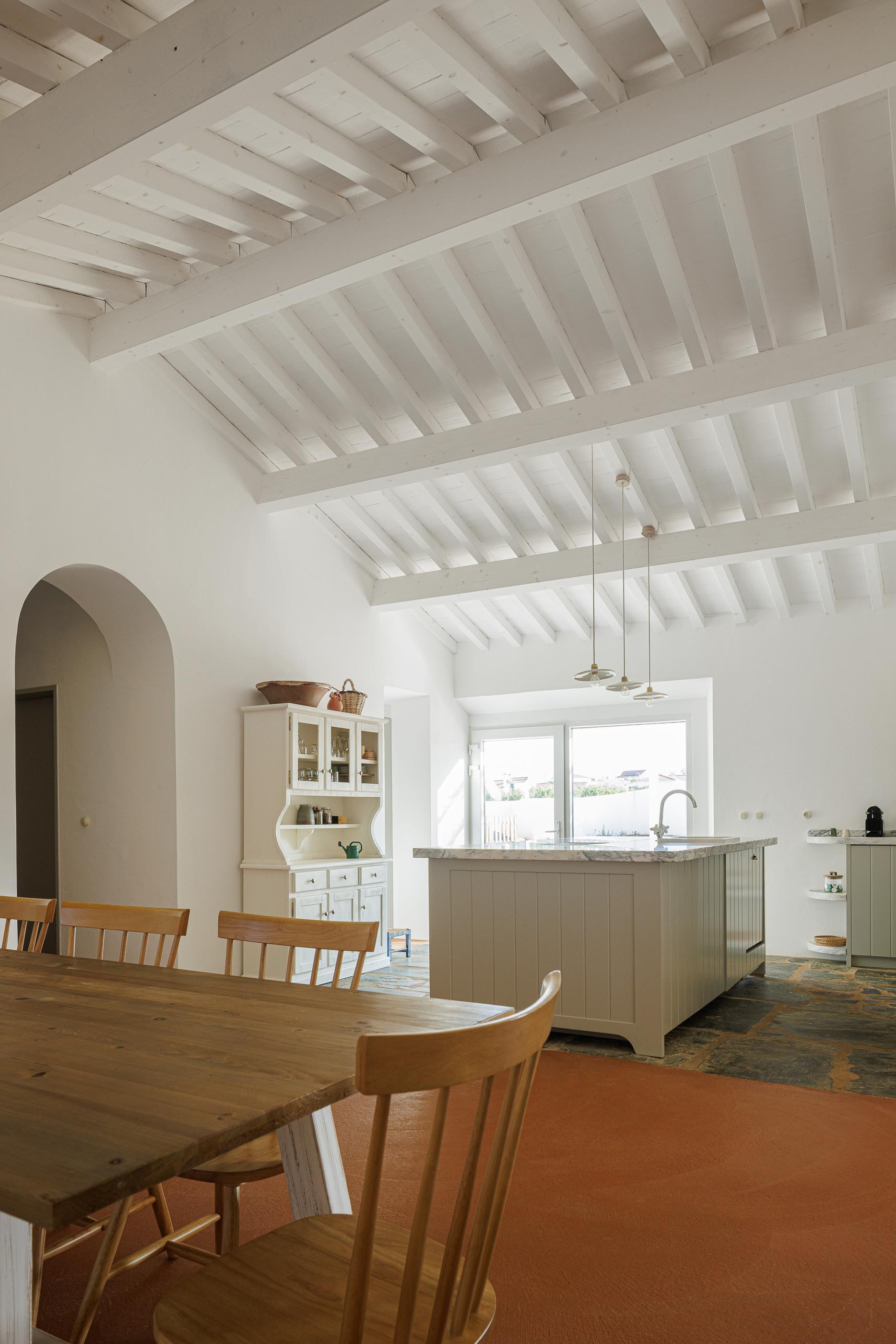 Cozinha/Sala de jantar: Vista geral com mesa de jantar, ilha de cozinha e teto branco com vigas de madeira. — Monte do Divor arquitetura Vasco Burnay Arquitectura em Estremoz