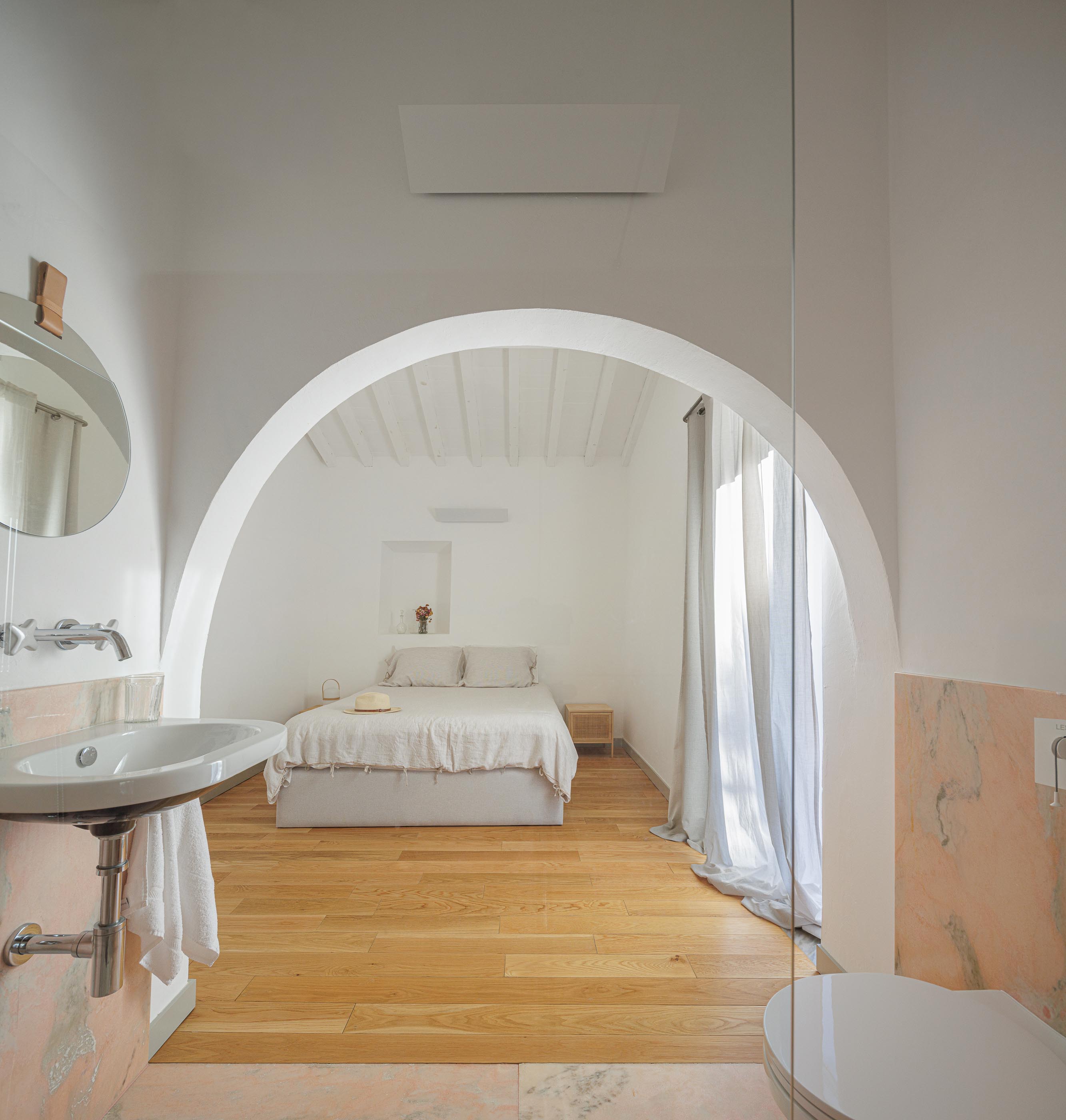 Casa de banho: Lavatório suspenso, espelho redondo e quarto com cama de casal ao fundo. — Monte do Divor arquitetura Vasco Burnay Arquitectura em Estremoz