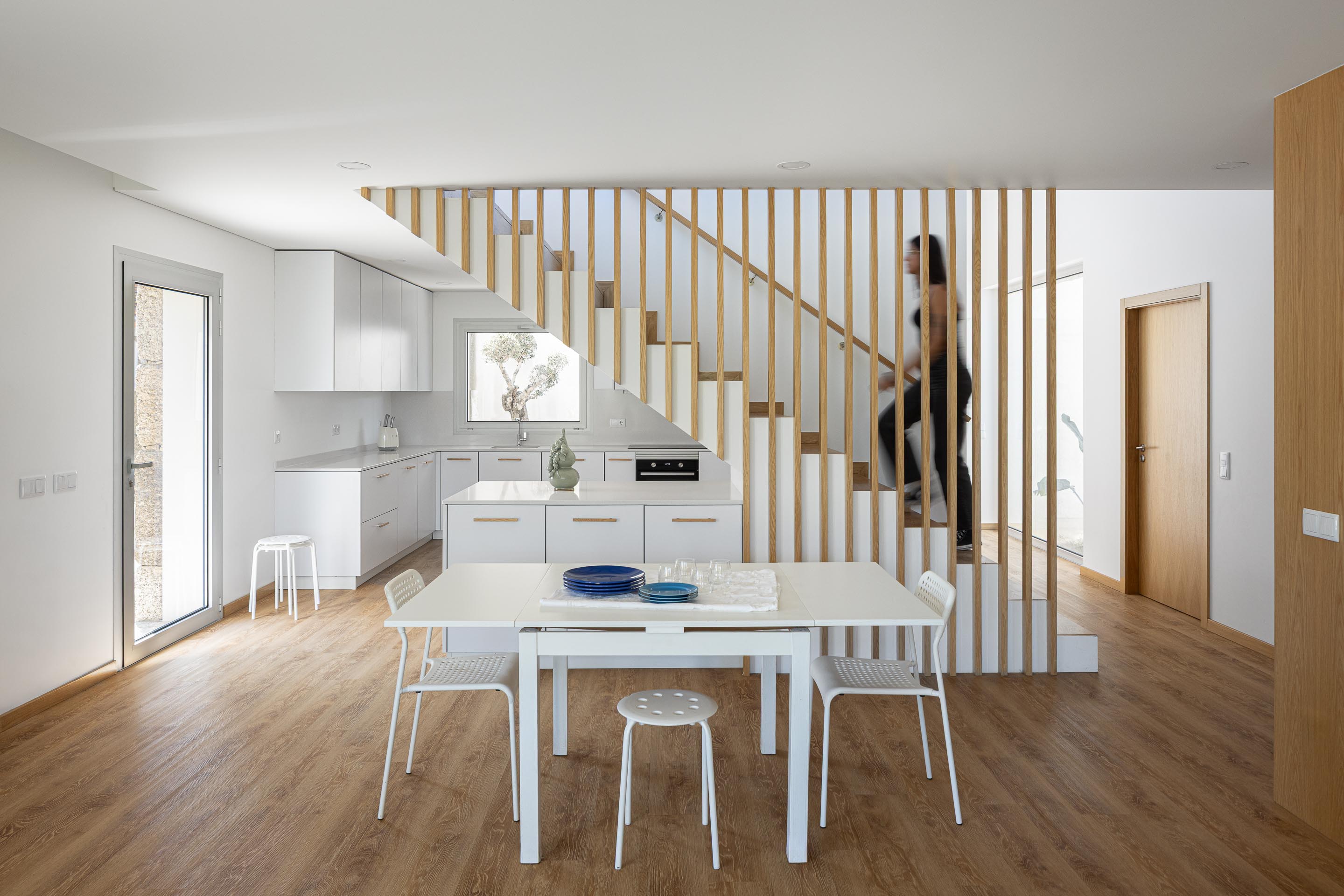 Cozinha/Sala de jantar: Espaço aberto com cozinha, mesa de jantar branca e escadas com ripas de madeira. — Casa em Mesão Frio arquitetura felixARQS – Daniel Félix Arquitectos em Guimarães