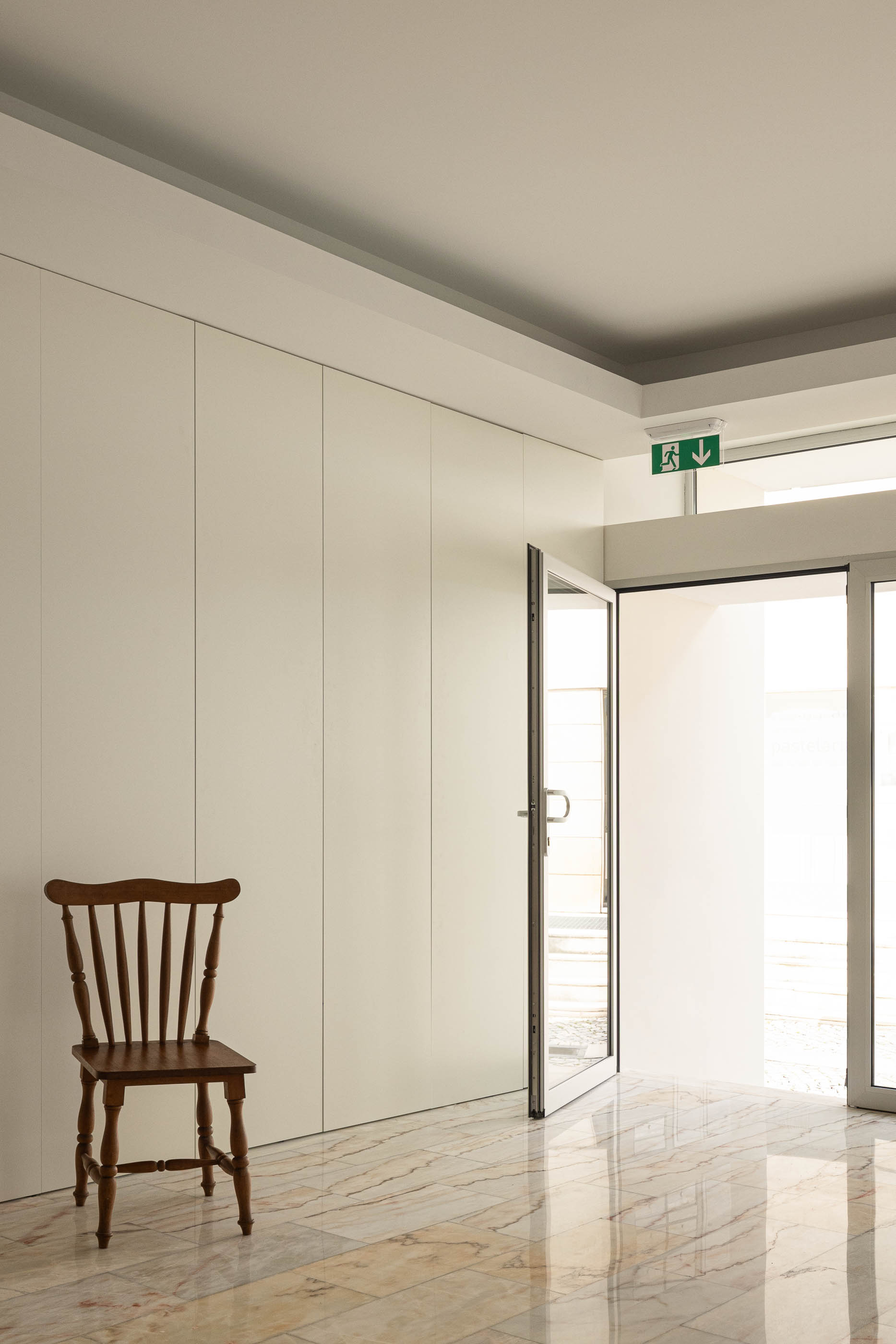 Hall de entrada com pavimento em mármore, paredes brancas e armários embutidos. Uma cadeira de madeira simples junto à porta de vidro aberta. — Reconversão do Talho Central arquitetura José Pedro Lima Arquiteto em Coimbra