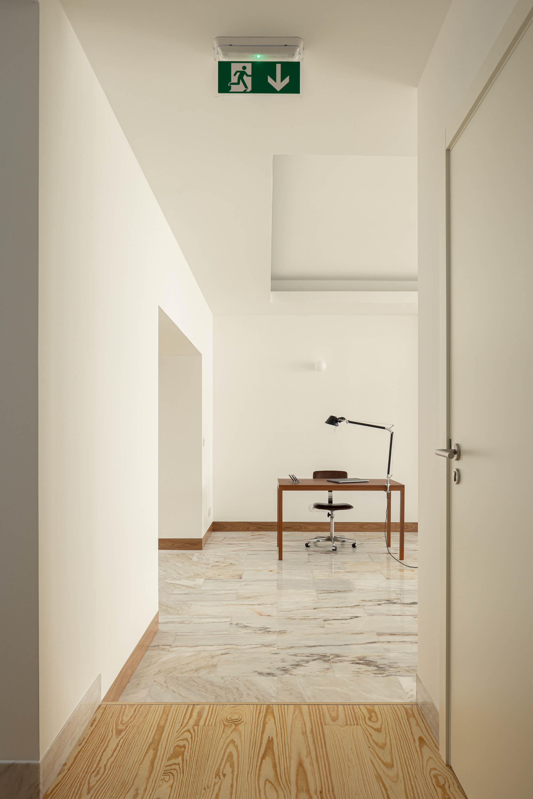 Corredor com pavimento em madeira que dá acesso a escritório com pavimento em mármore. Sinalética de emergência no teto branco. — Reconversão do Talho Central arquitetura José Pedro Lima Arquiteto em Coimbra