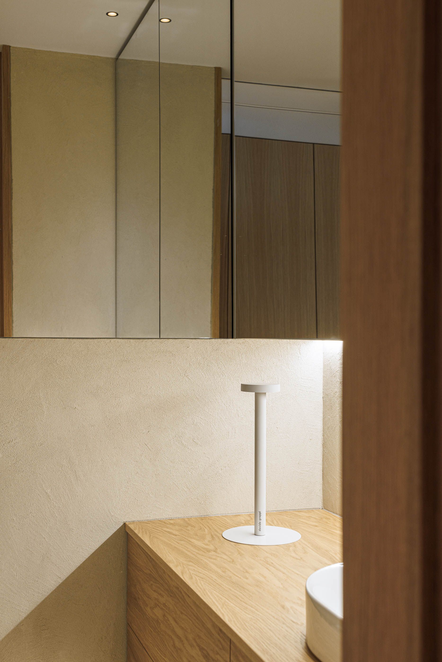 Casa de banho: Detalhe do lavatório sobre móvel em madeira clara, espelho e candeeiro de mesa minimalista. — Barra Apartment arquitetura Paulo Martins Arquitectura e Design em Aveiro