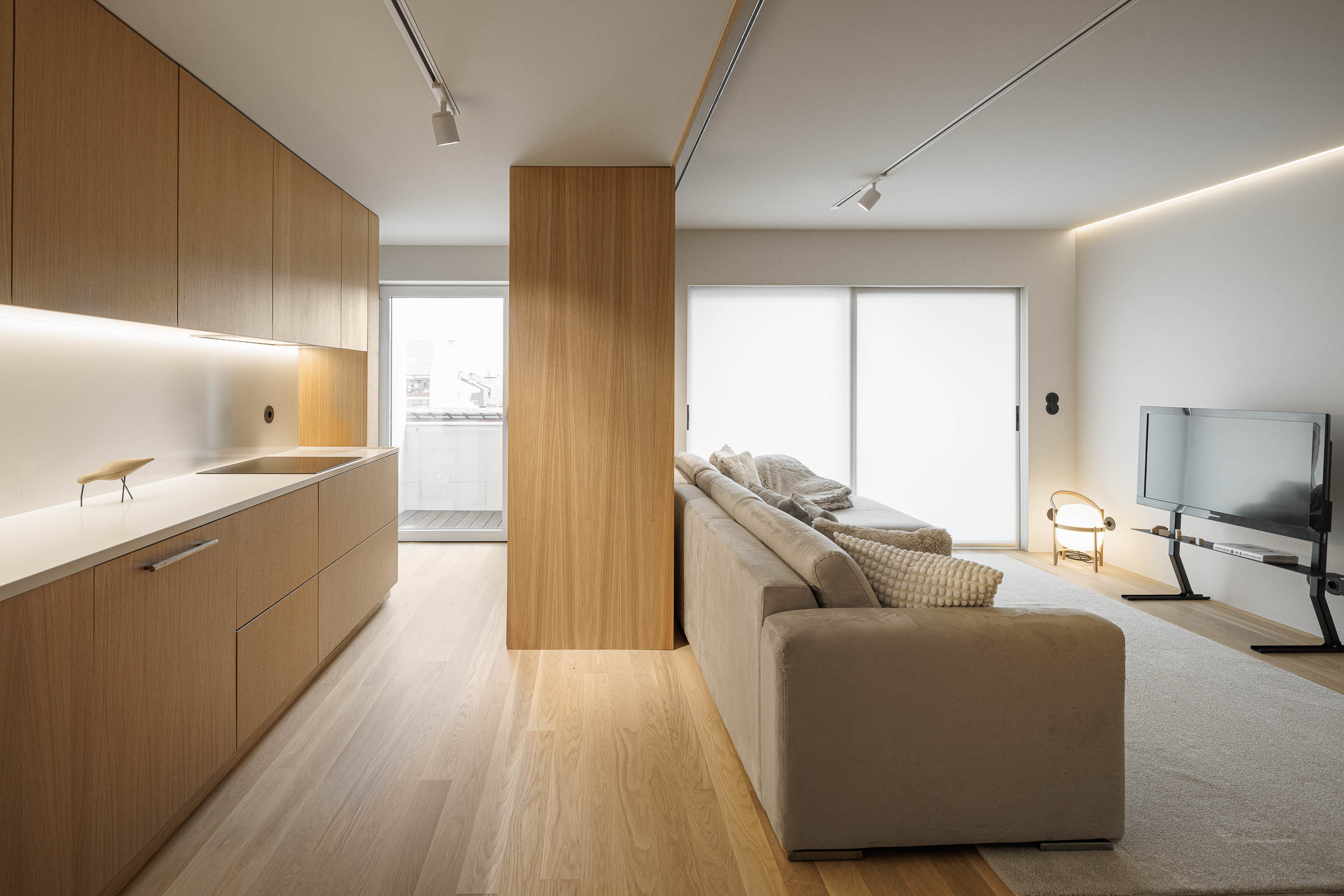 Sala de estar: Cozinha em madeira clara à esquerda, sofá bege e televisão na parede, com alcatifa clara. — Barra Apartment arquitetura Paulo Martins Arquitectura e Design em Aveiro
