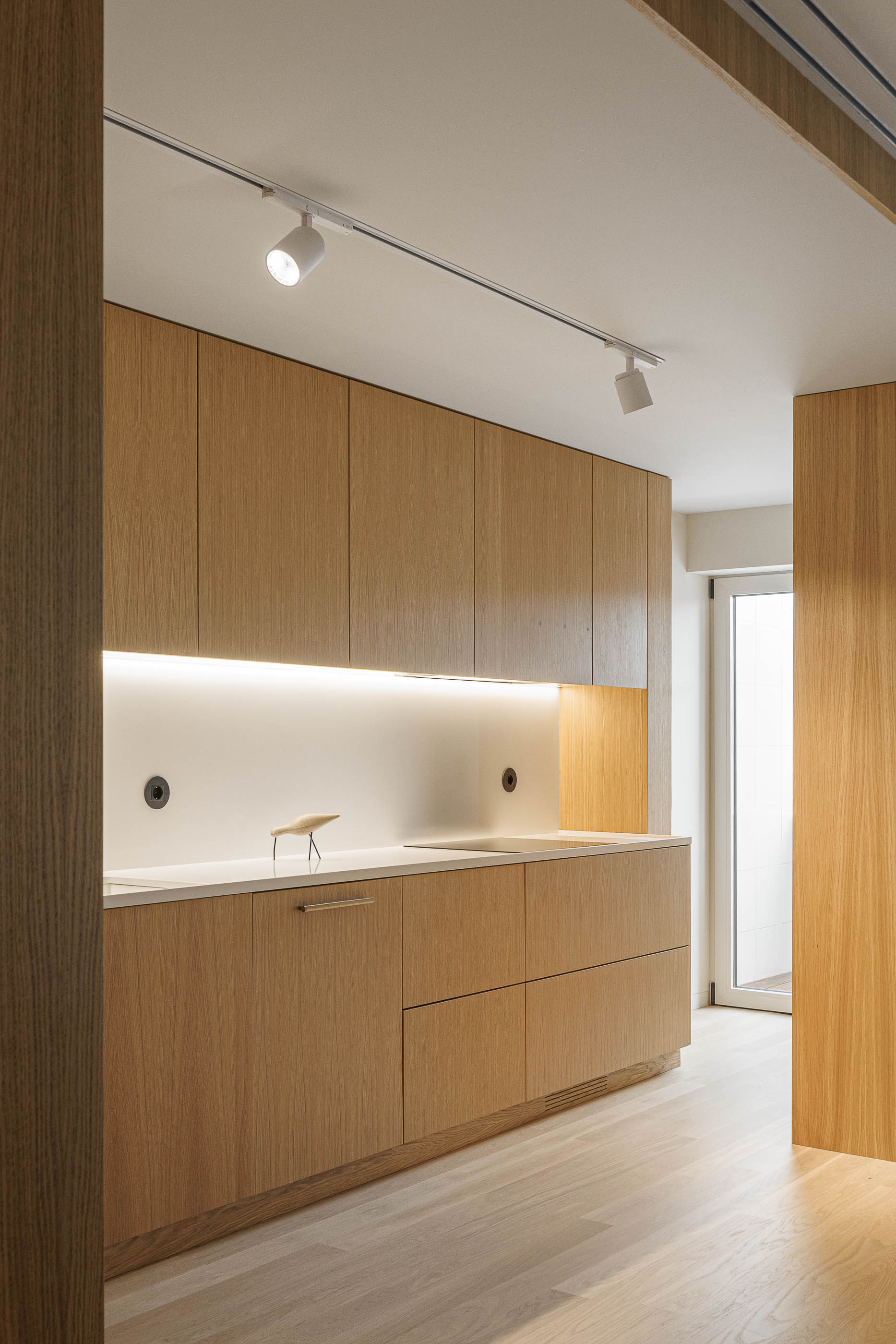 Cozinha: Armários em madeira clara com iluminação embutida e pavimento em madeira, com candeeiro de teto. — Barra Apartment arquitetura Paulo Martins Arquitectura e Design em Aveiro