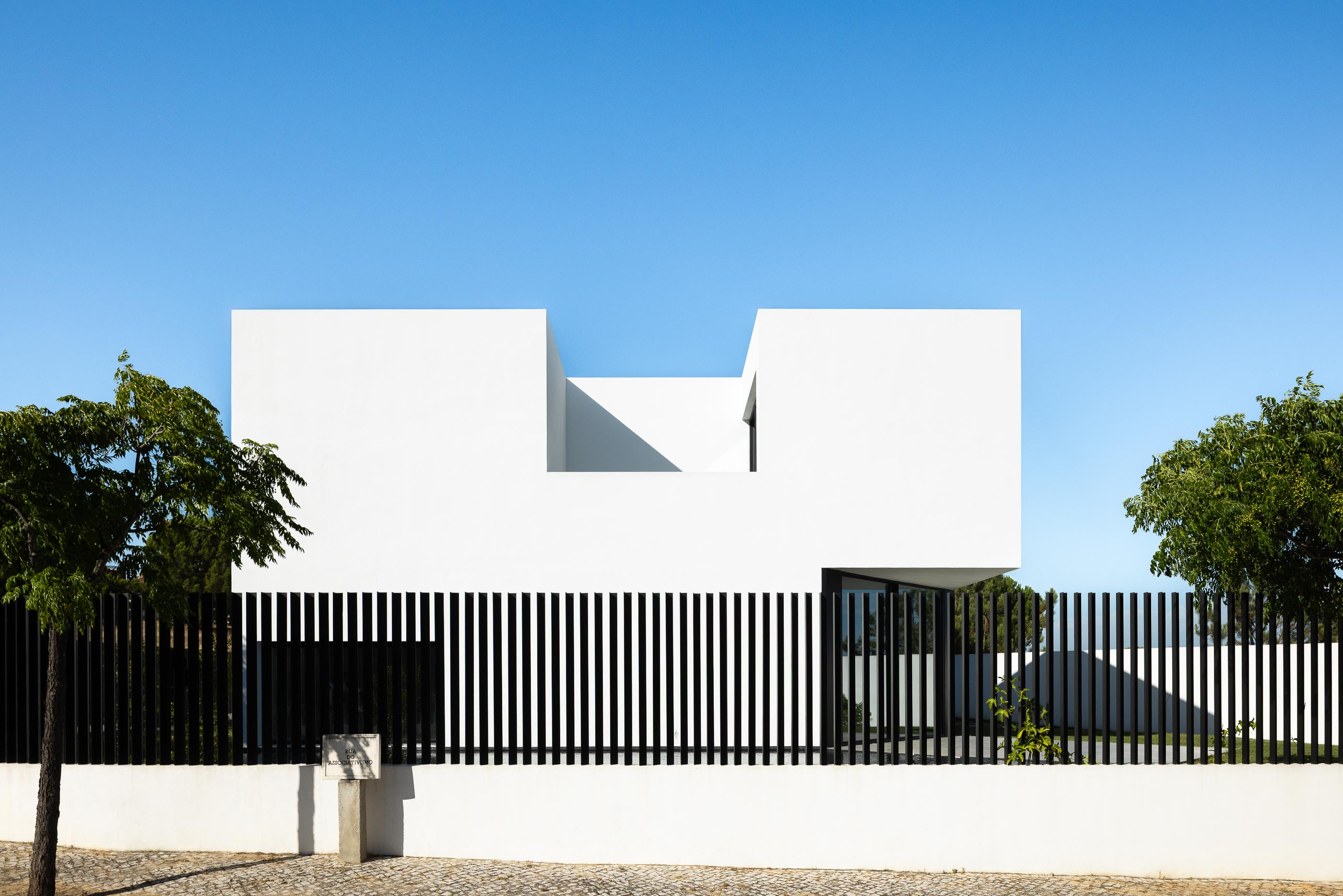 Fachada: Vista frontal da casa minimalista branca com vedação preta vertical e árvores. — Casa DR arquitetura NMDARQ – Nuno Miguel Dias, Arquitecto em Torres Novas