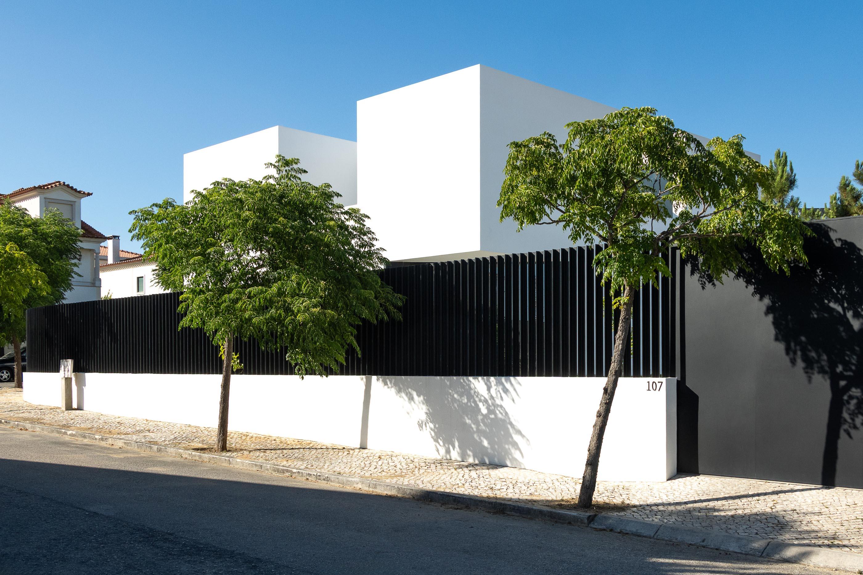 Fachada: Perspectiva angular da casa branca com vedação preta, árvore e número 107. — Casa DR arquitetura NMDARQ – Nuno Miguel Dias, Arquitecto em Torres Novas