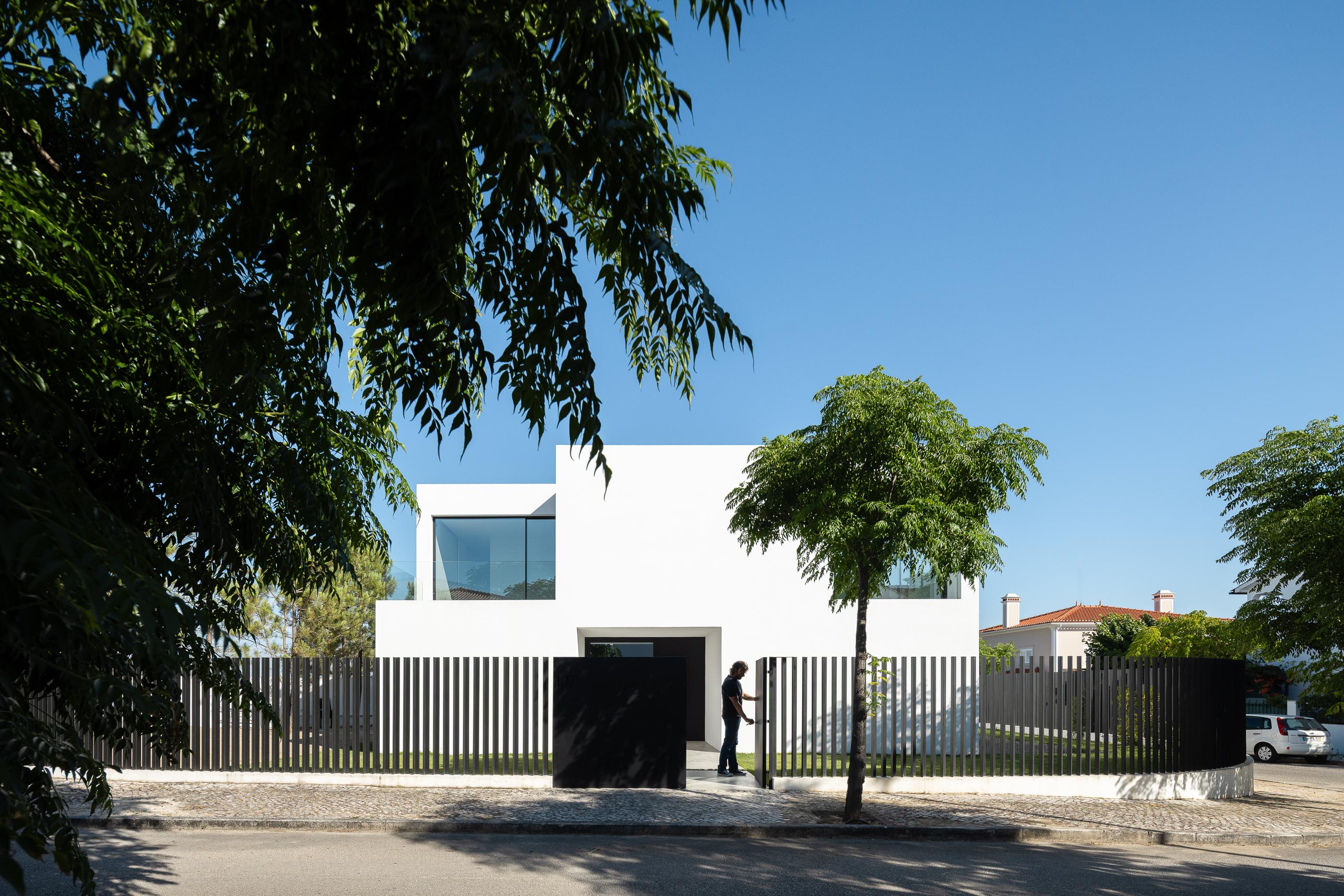 Fachada: Casa branca minimalista com vedação preta, pessoa a entrar e vegetação. — Casa DR arquitetura NMDARQ – Nuno Miguel Dias, Arquitecto em Torres Novas