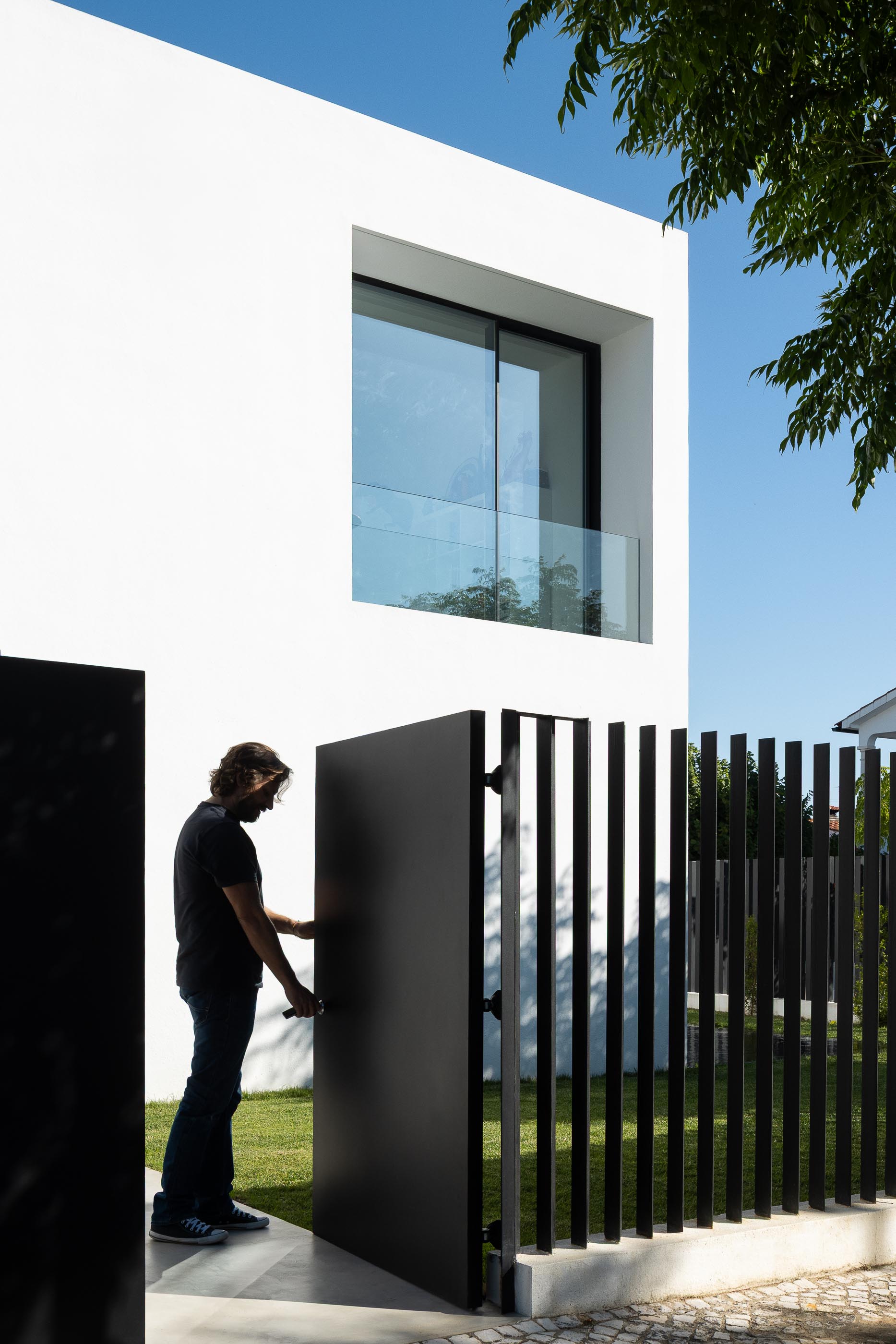 Exterior: Homem a abrir portão preto da vedação da casa branca minimalista. — Casa DR arquitetura NMDARQ – Nuno Miguel Dias, Arquitecto em Torres Novas