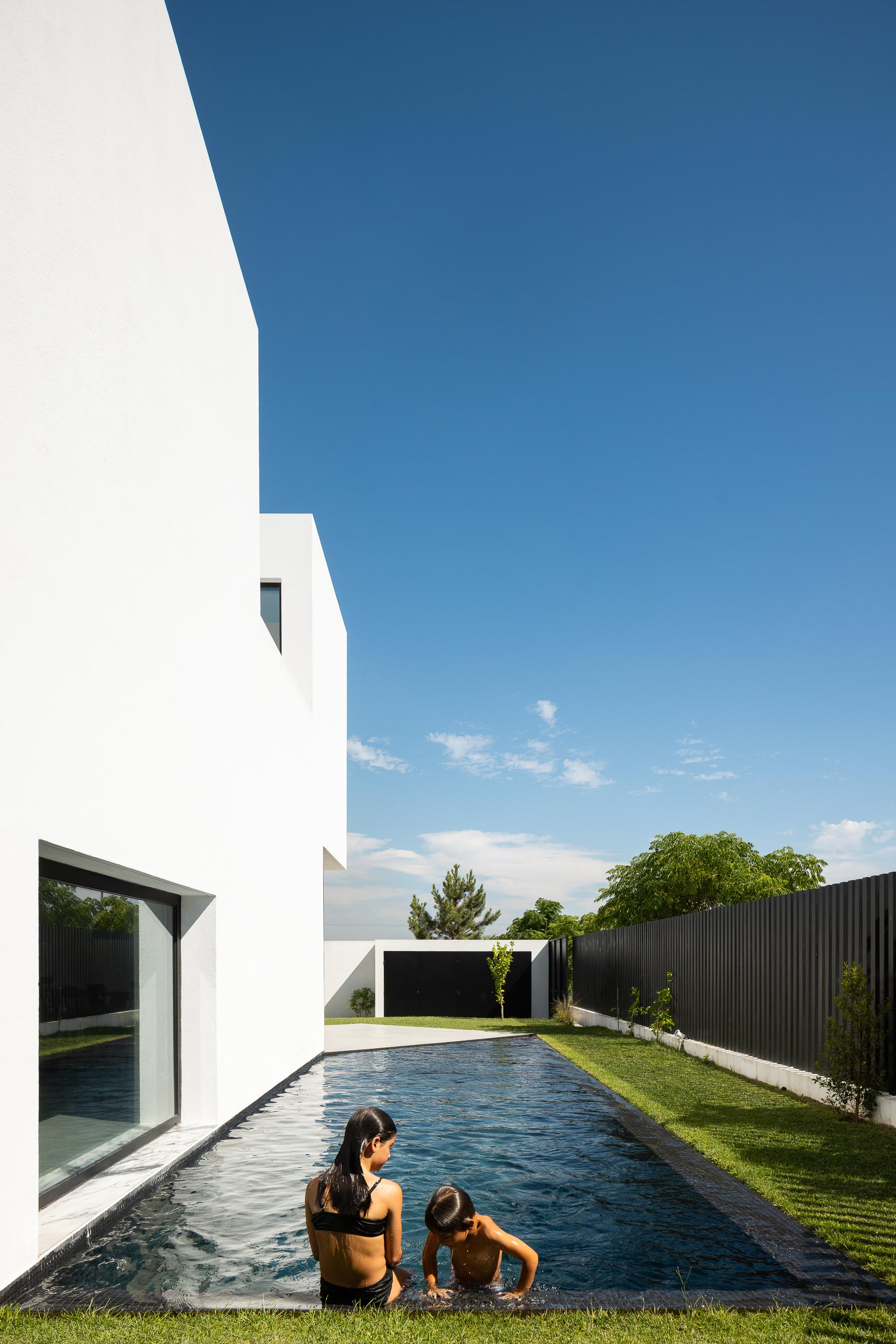 Interior: Criança na piscina vista do interior, com cadeiras pretas e pavimento cinza. — Casa DR arquitetura NMDARQ – Nuno Miguel Dias, Arquitecto em Torres Novas