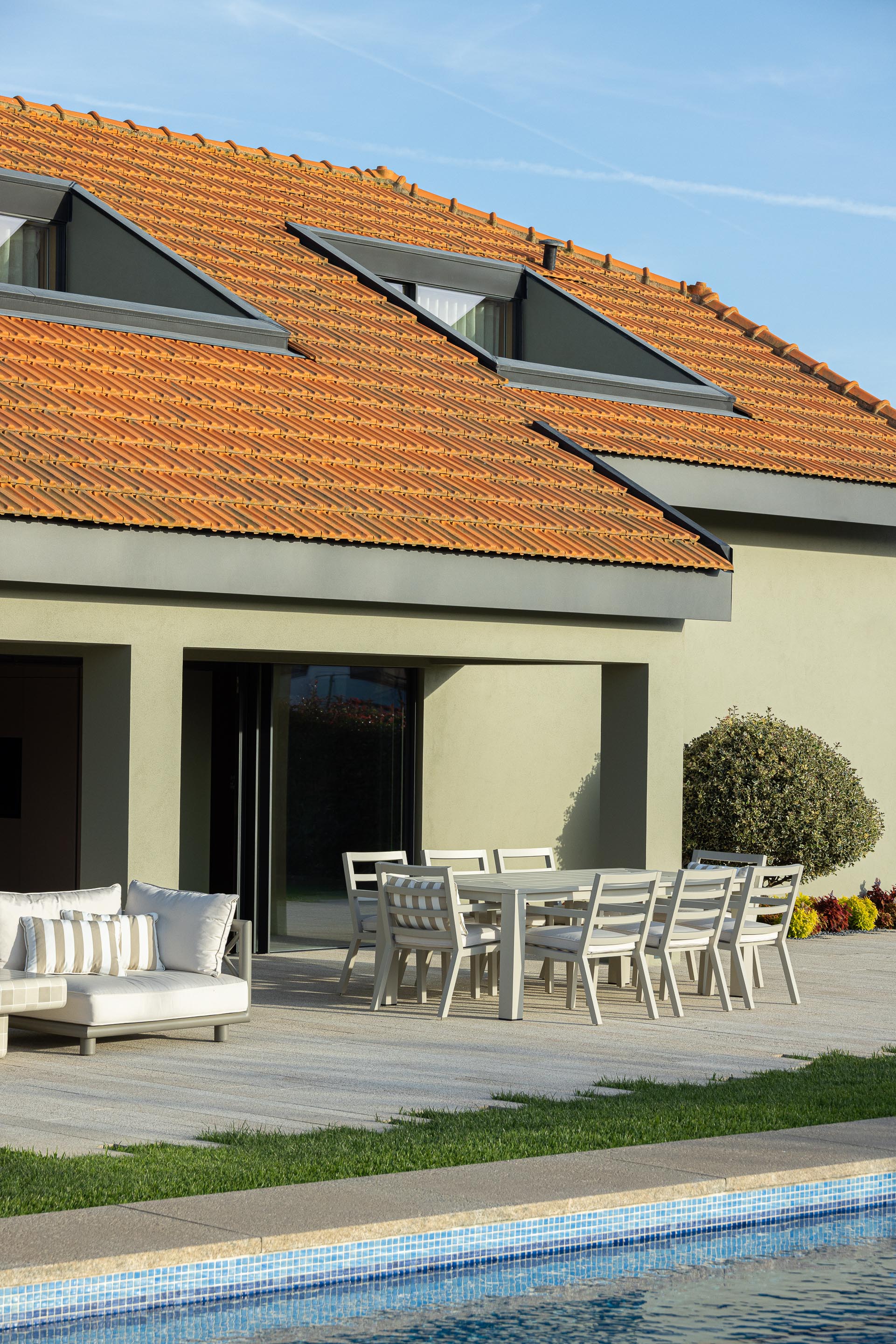 Terraço: Mesa de jantar exterior em pedra clara, cadeiras brancas e sofás com almofadas listradas, junto à piscina. Telhado de telha tradicional. — Casa do Engenho arquitetura silverline em Vila Nova de Gaia