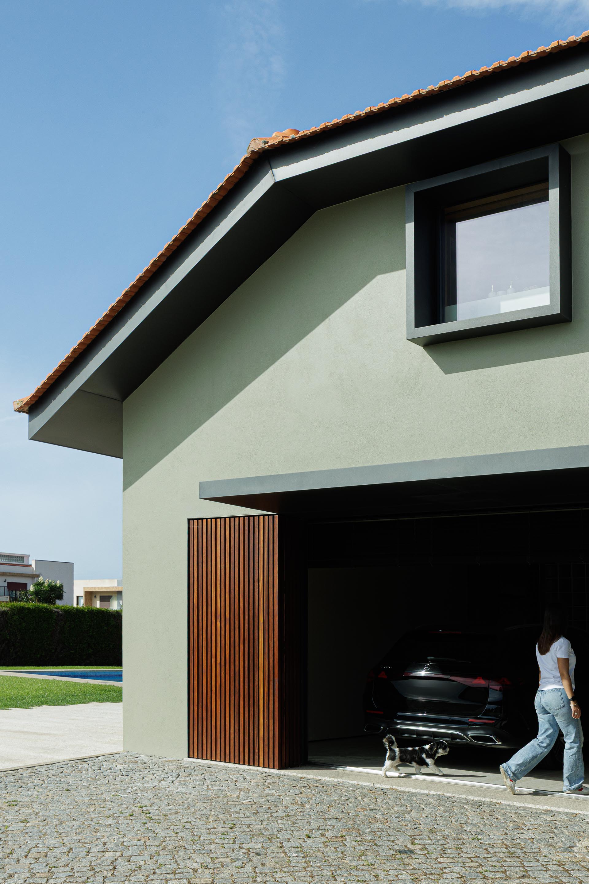 Garagem: Fachada com porta em ripas de madeira parcialmente aberta, revelando um carro e uma pessoa. Pavimento em calçada. — Casa do Engenho arquitetura silverline em Vila Nova de Gaia