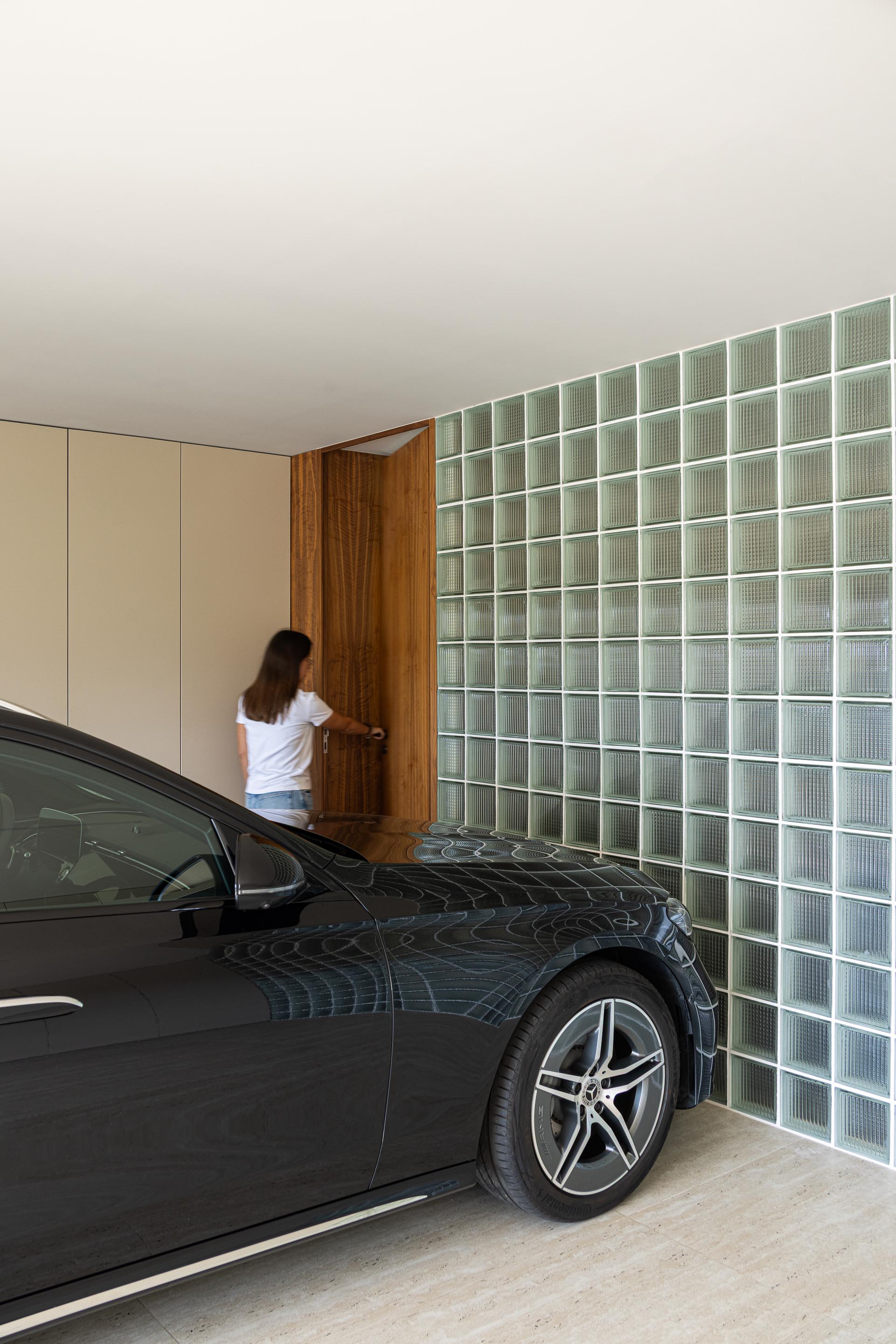 Garagem: Interior com parede de blocos de vidro, porta em madeira e um carro preto estacionado. — Casa do Engenho arquitetura silverline em Vila Nova de Gaia
