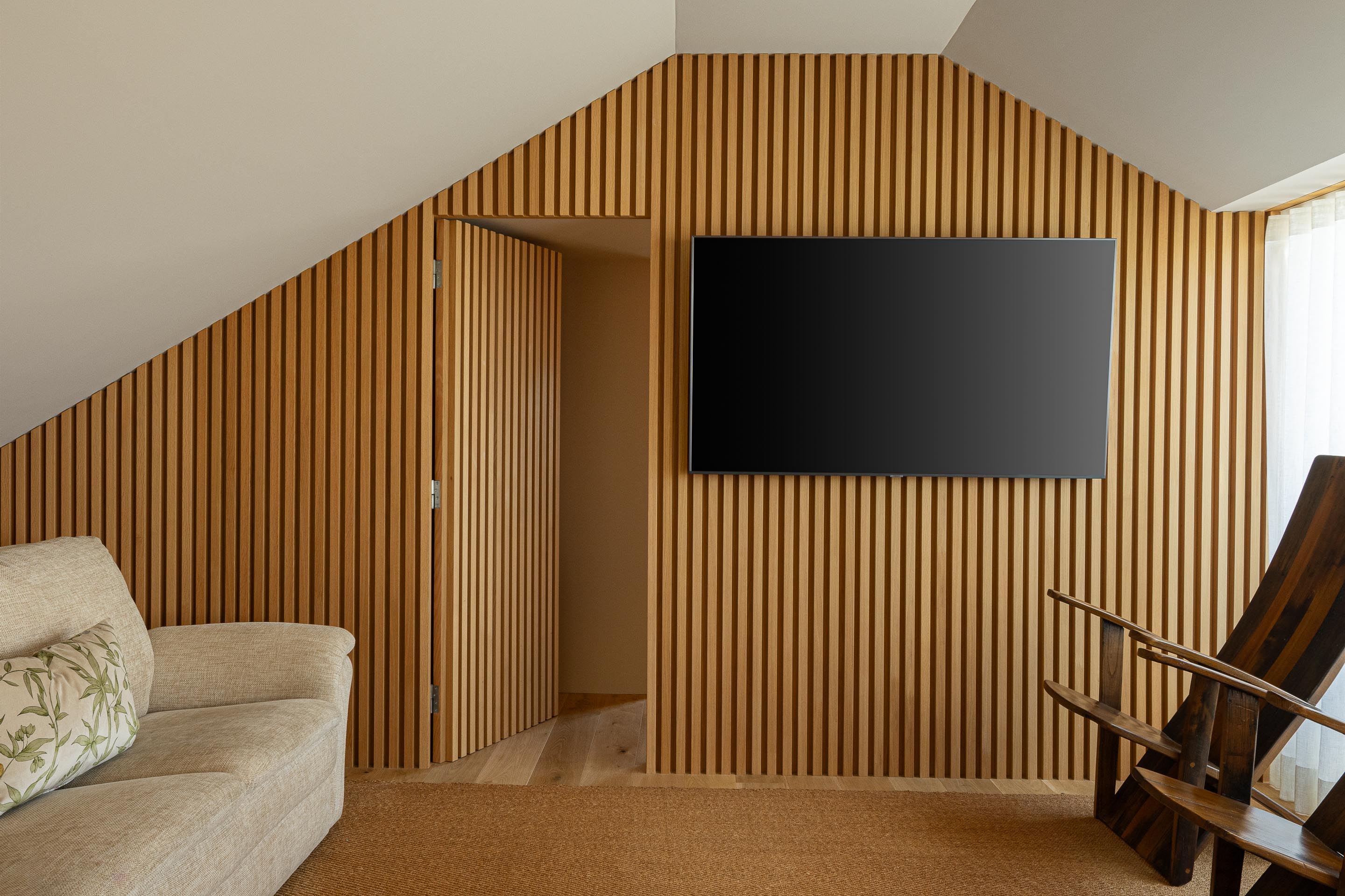 Sala de estar: Parede revestida em ripas de madeira clara, televisão, sofá bege e cadeiras de madeira escura. — Casa do Engenho arquitetura silverline em Vila Nova de Gaia
