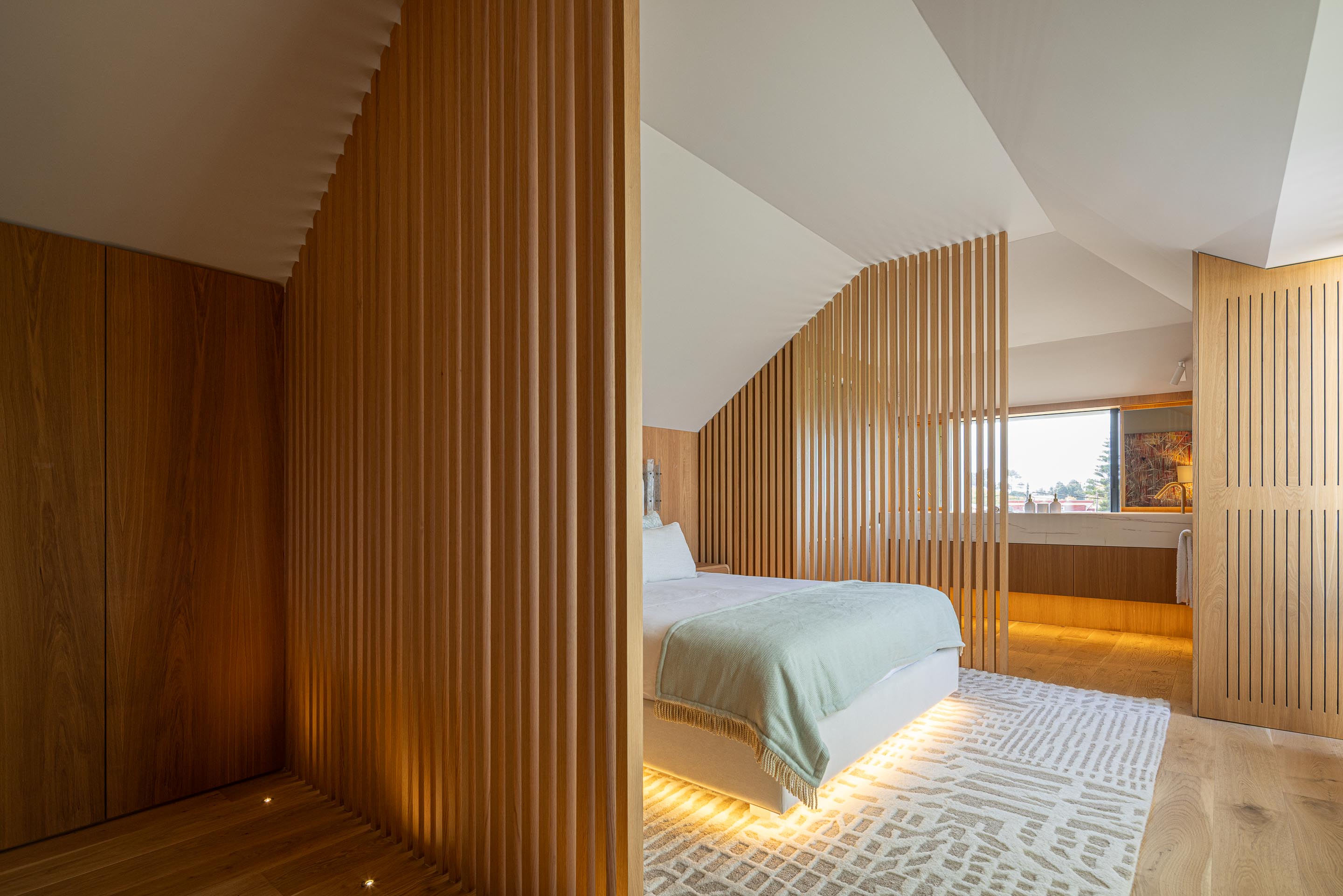 Quarto: Divisória em ripas de madeira clara, cama com roupa de cama verde clara, alcatifa geométrica e casa de banho ao fundo. — Casa do Engenho arquitetura silverline em Vila Nova de Gaia