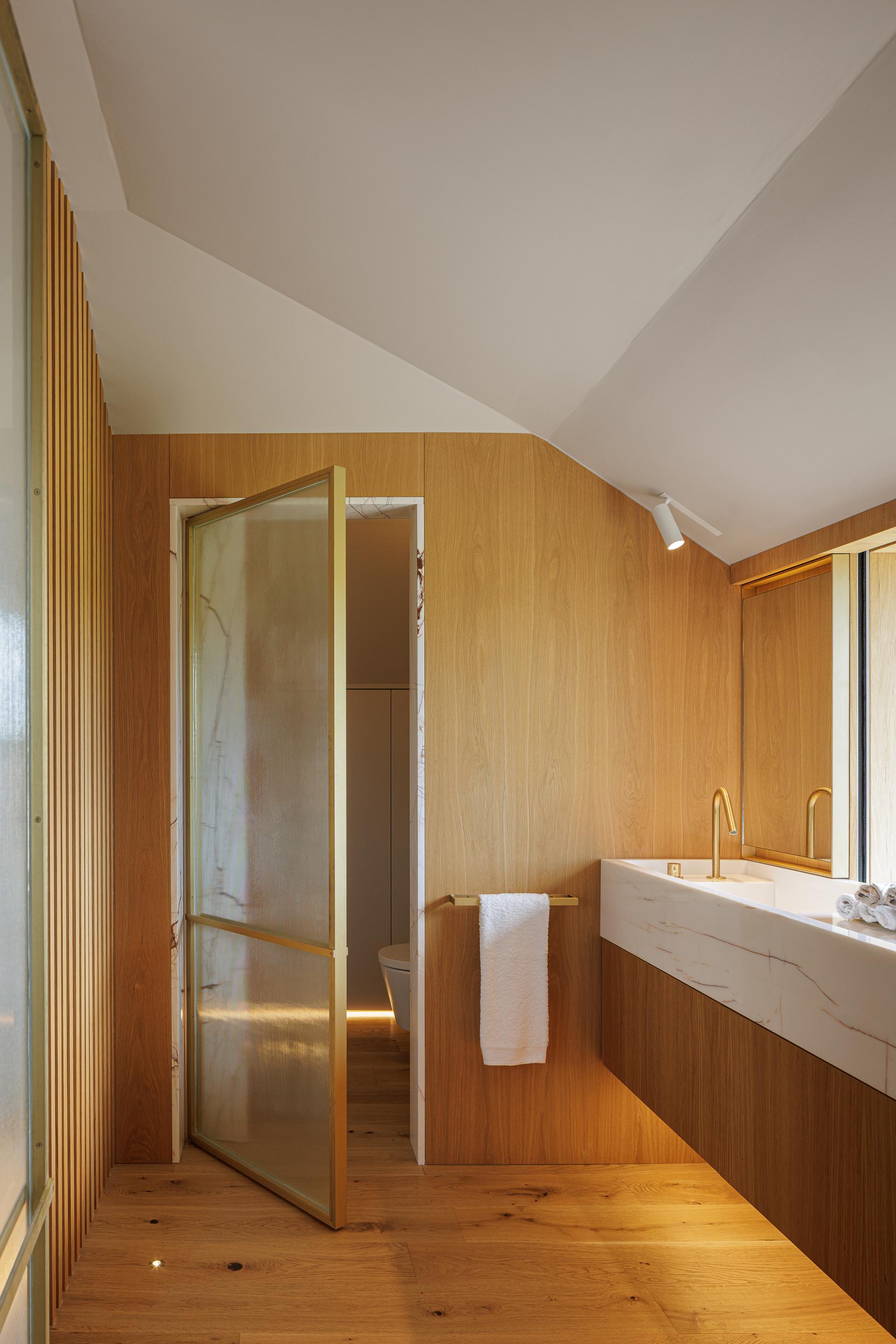 Casa de banho: Lavatório em mármore branco, torneira dourada, cabine de duche com porta em vidro e parede revestida em madeira clara. — Casa do Engenho arquitetura silverline em Vila Nova de Gaia