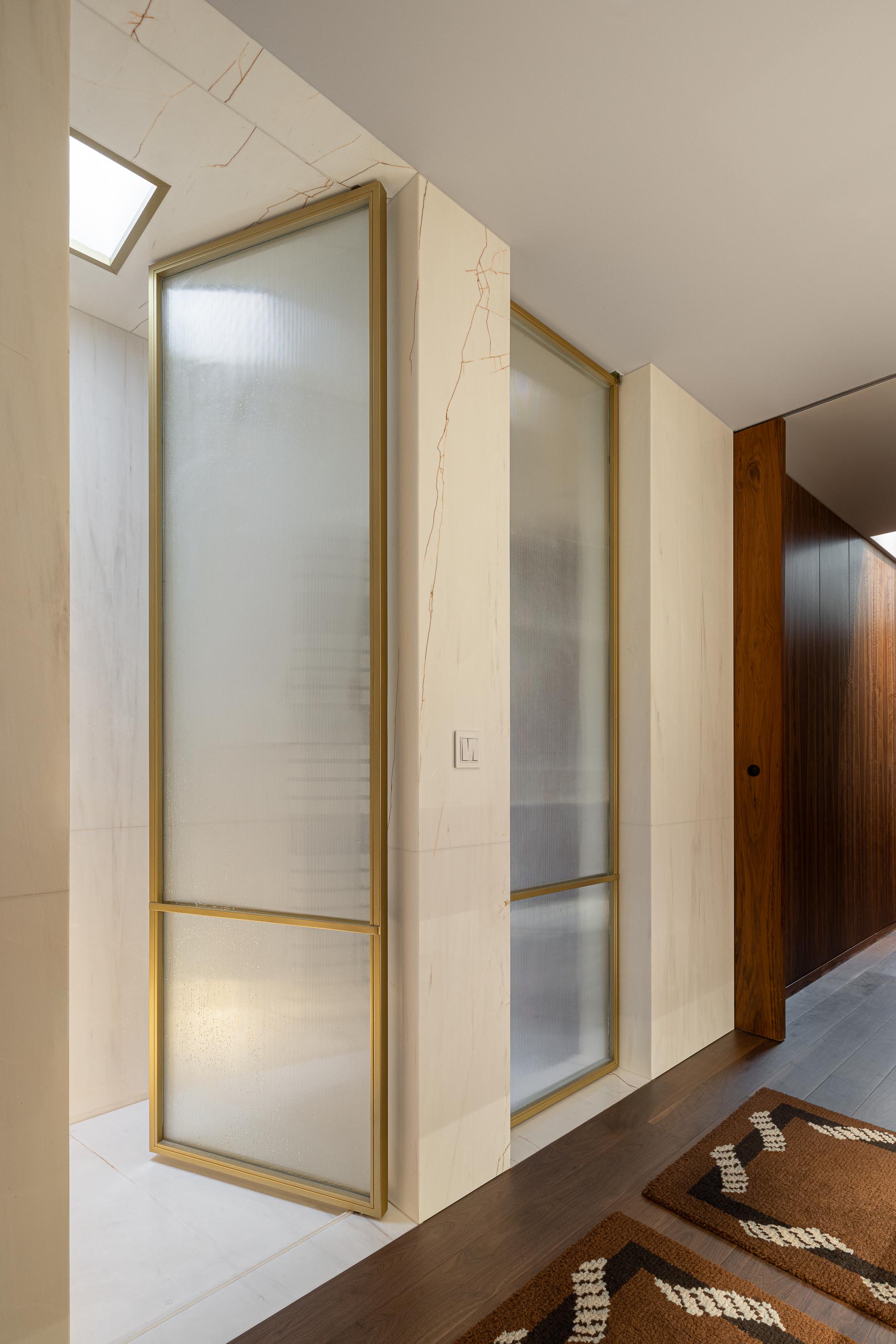 Casa de banho: Cabine de duche com portas em vidro canelado e estrutura dourada, paredes em mármore e tapetes geométricos. — Casa do Engenho arquitetura silverline em Vila Nova de Gaia