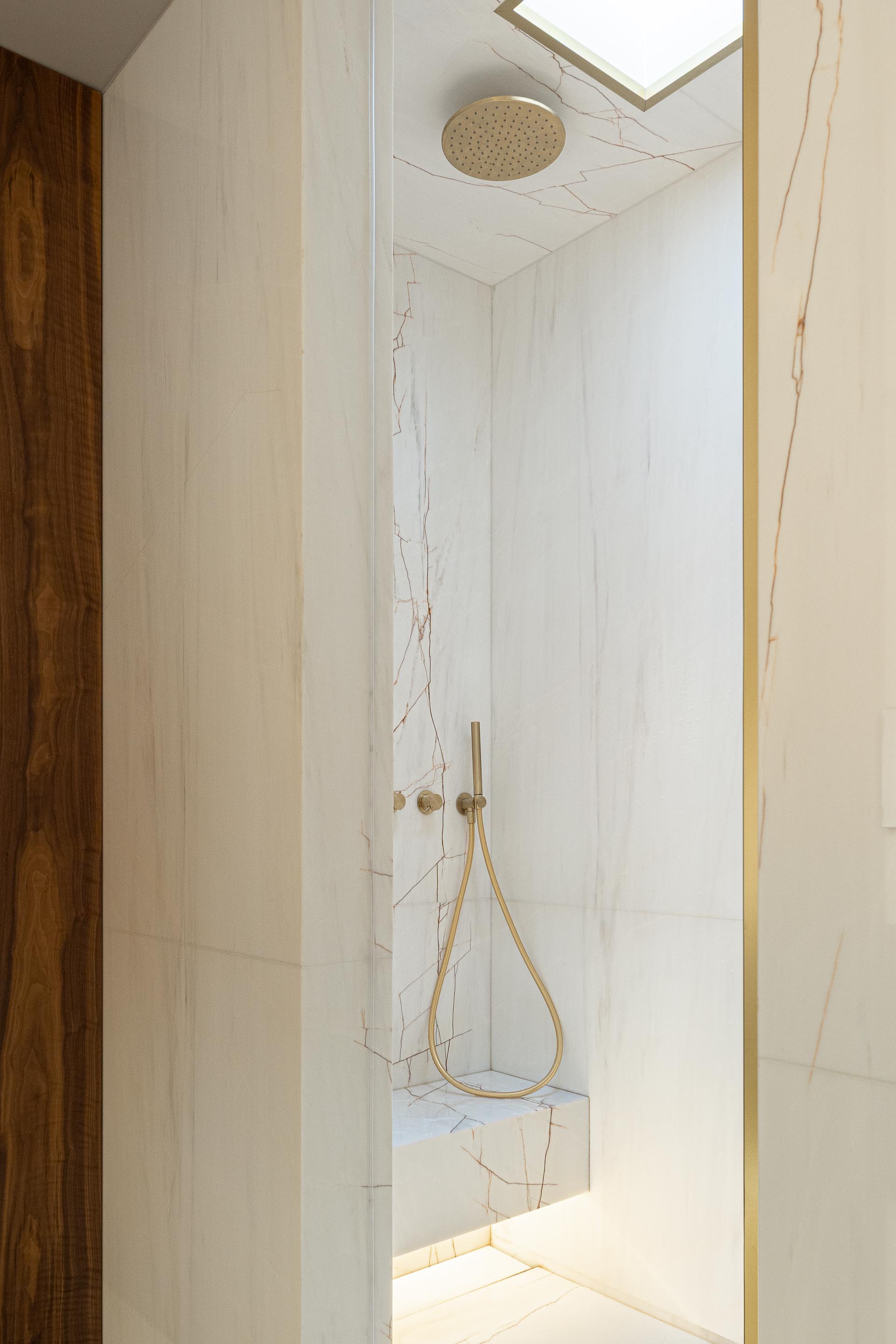 Casa de banho: Interior da cabine de duche com revestimento em mármore branco, chuveiro dourado e banco iluminado. — Casa do Engenho arquitetura silverline em Vila Nova de Gaia