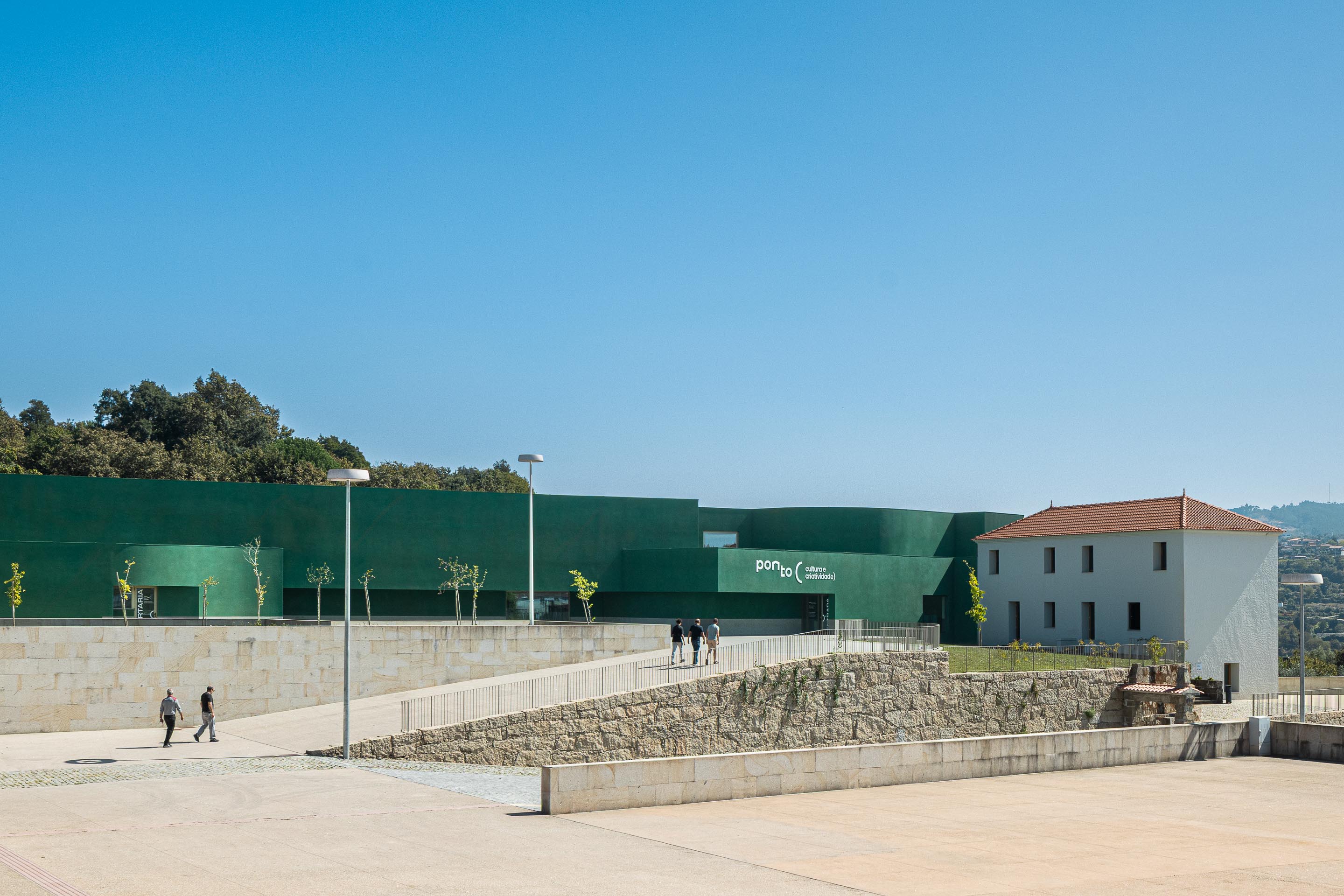 Fachada: Vista exterior do edifício verde "Ponto C" com acesso pedonal e muro de pedra. — Ponto C - Cultura e Criatividade arquitetura Galeria Gabinete em Penafiel