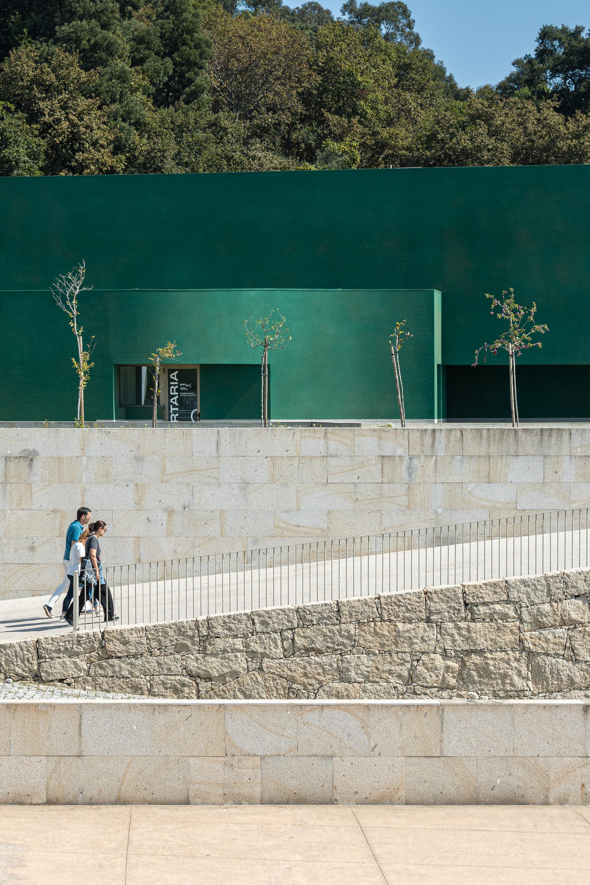 Exterior: Rampa de acesso ao edifício verde, com parede de pedra e sinalética da receção. — Ponto C - Cultura e Criatividade arquitetura Galeria Gabinete em Penafiel