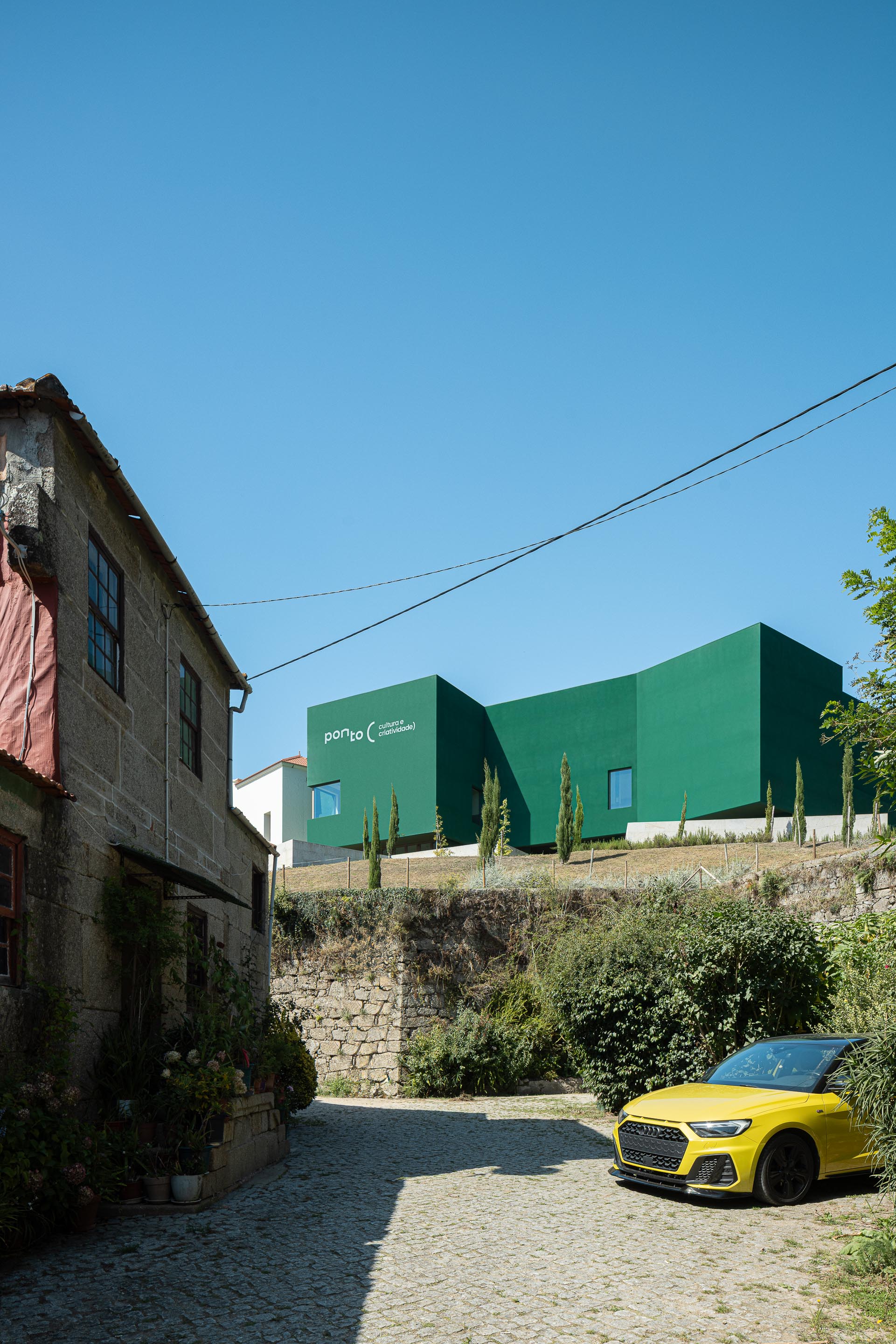 Exterior: Edifício verde "Ponto C" visto de uma rua de calçada, com casa antiga em pedra. — Ponto C - Cultura e Criatividade arquitetura Galeria Gabinete em Penafiel