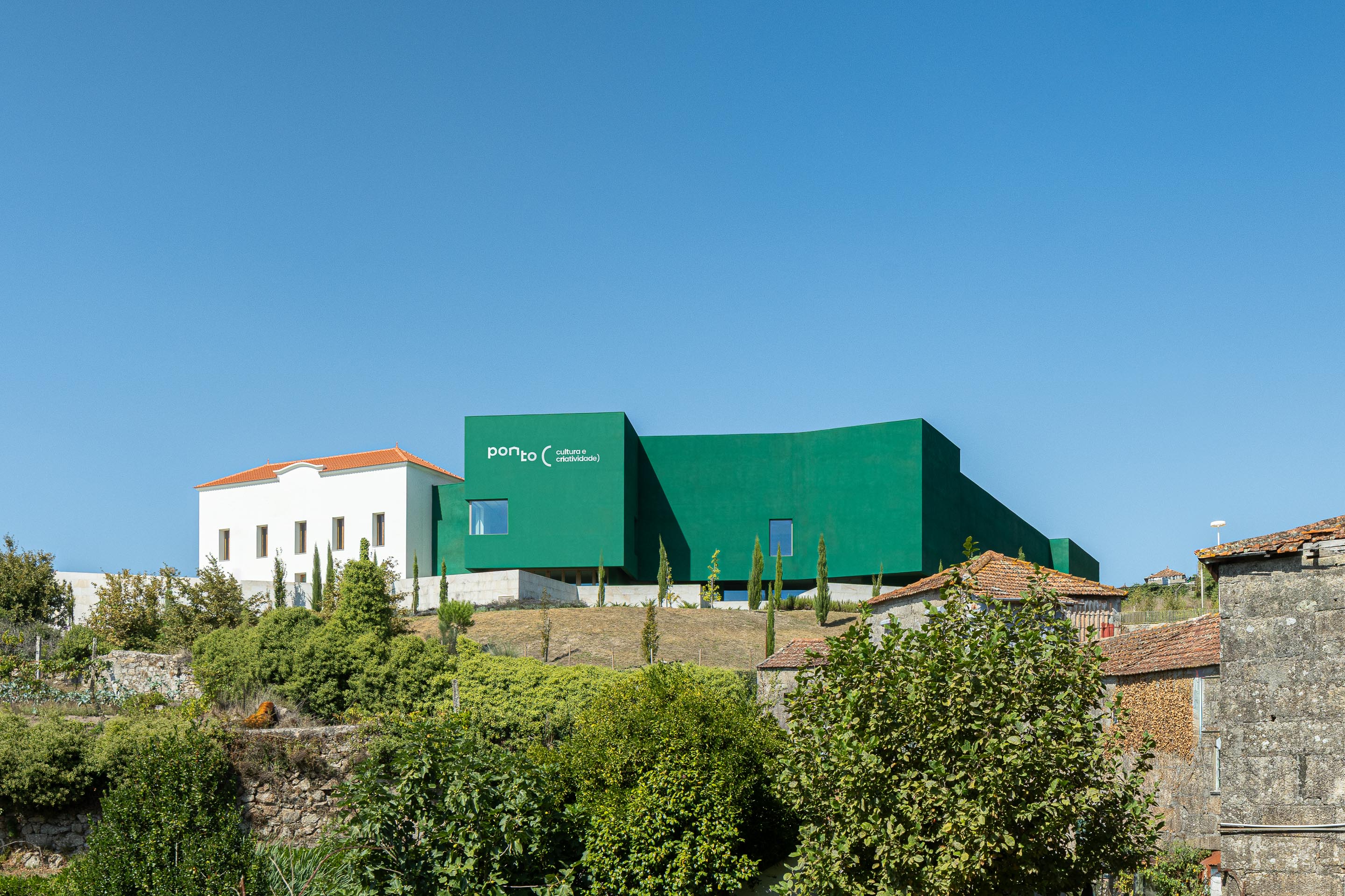 Exterior: Vista do edifício verde "Ponto C" no topo de uma colina, com vegetação e casas antigas. — Ponto C - Cultura e Criatividade arquitetura Galeria Gabinete em Penafiel