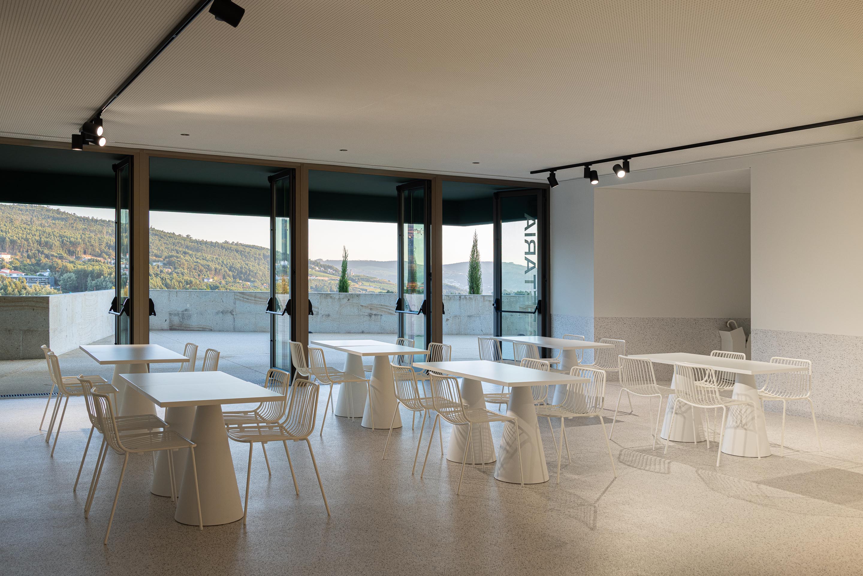 Cafetaria: Mesas e cadeiras brancas com vista para a paisagem através de janelas amplas. — Ponto C - Cultura e Criatividade arquitetura Galeria Gabinete em Penafiel
