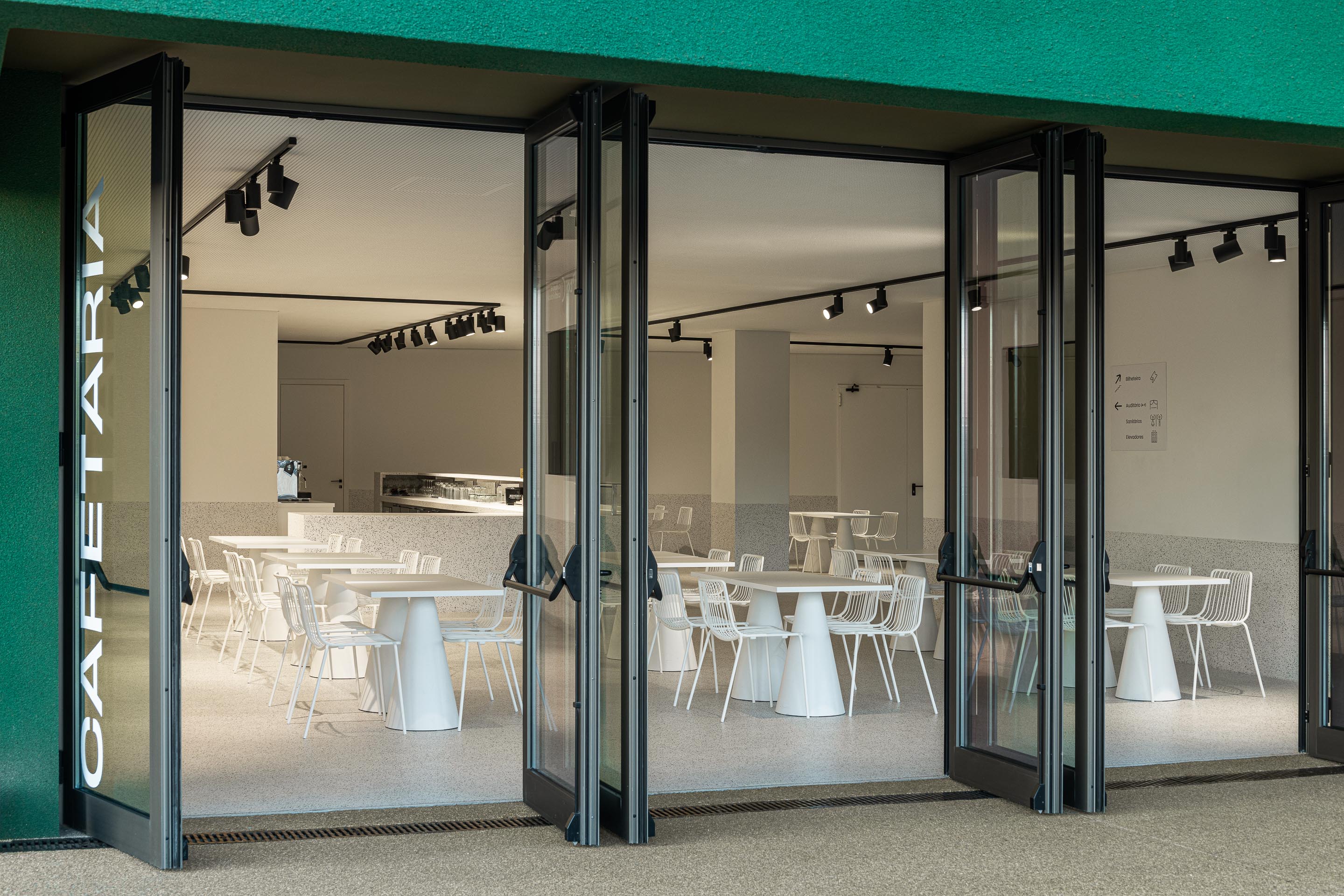 Cafetaria: Vista interior da cafetaria com mesas e cadeiras brancas através das portas de vidro. — Ponto C - Cultura e Criatividade arquitetura Galeria Gabinete em Penafiel