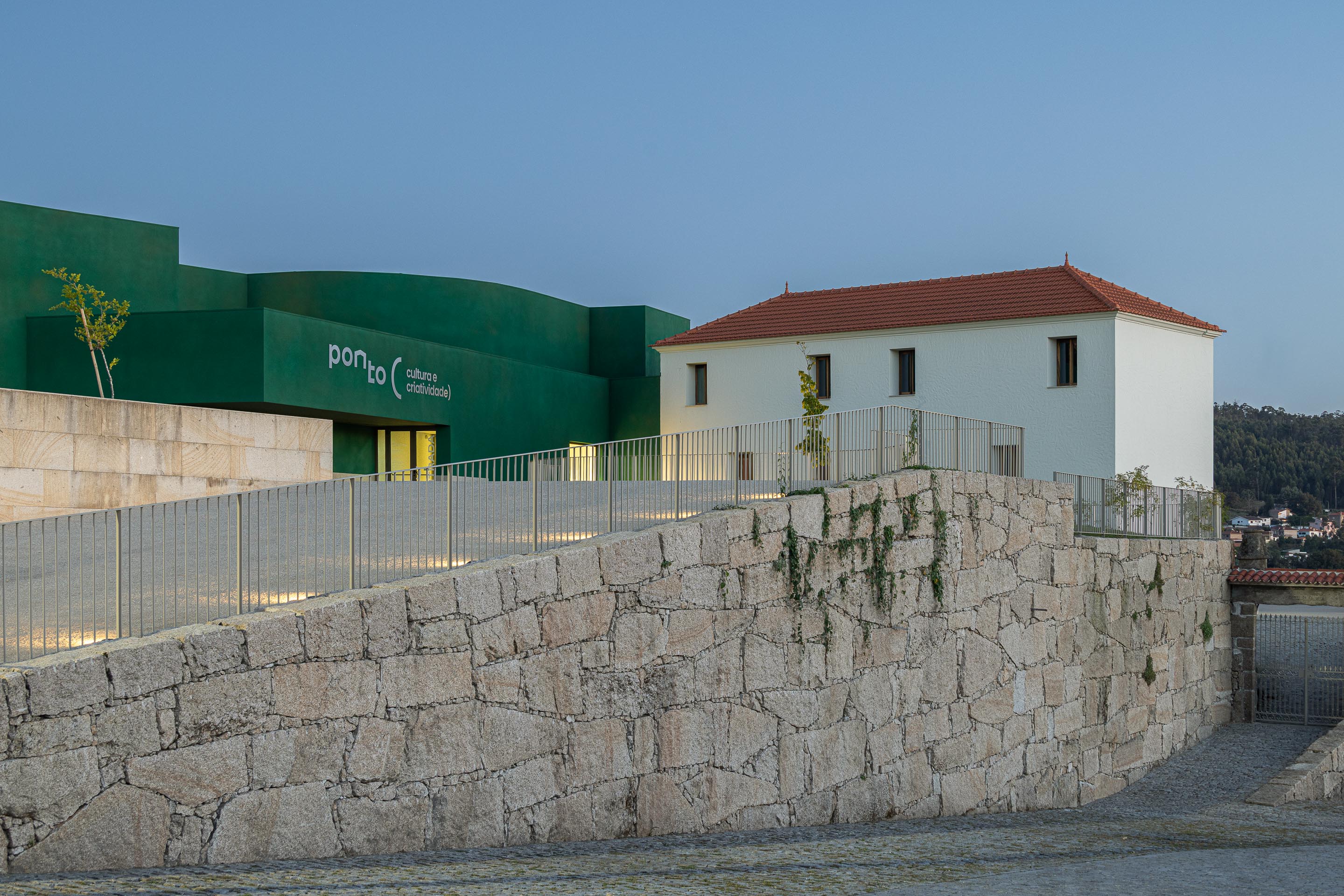 Fachada: Vista noturna do edifício verde "Ponto C" e edifício branco ao lado, com muro de pedra. — Ponto C - Cultura e Criatividade arquitetura Galeria Gabinete em Penafiel