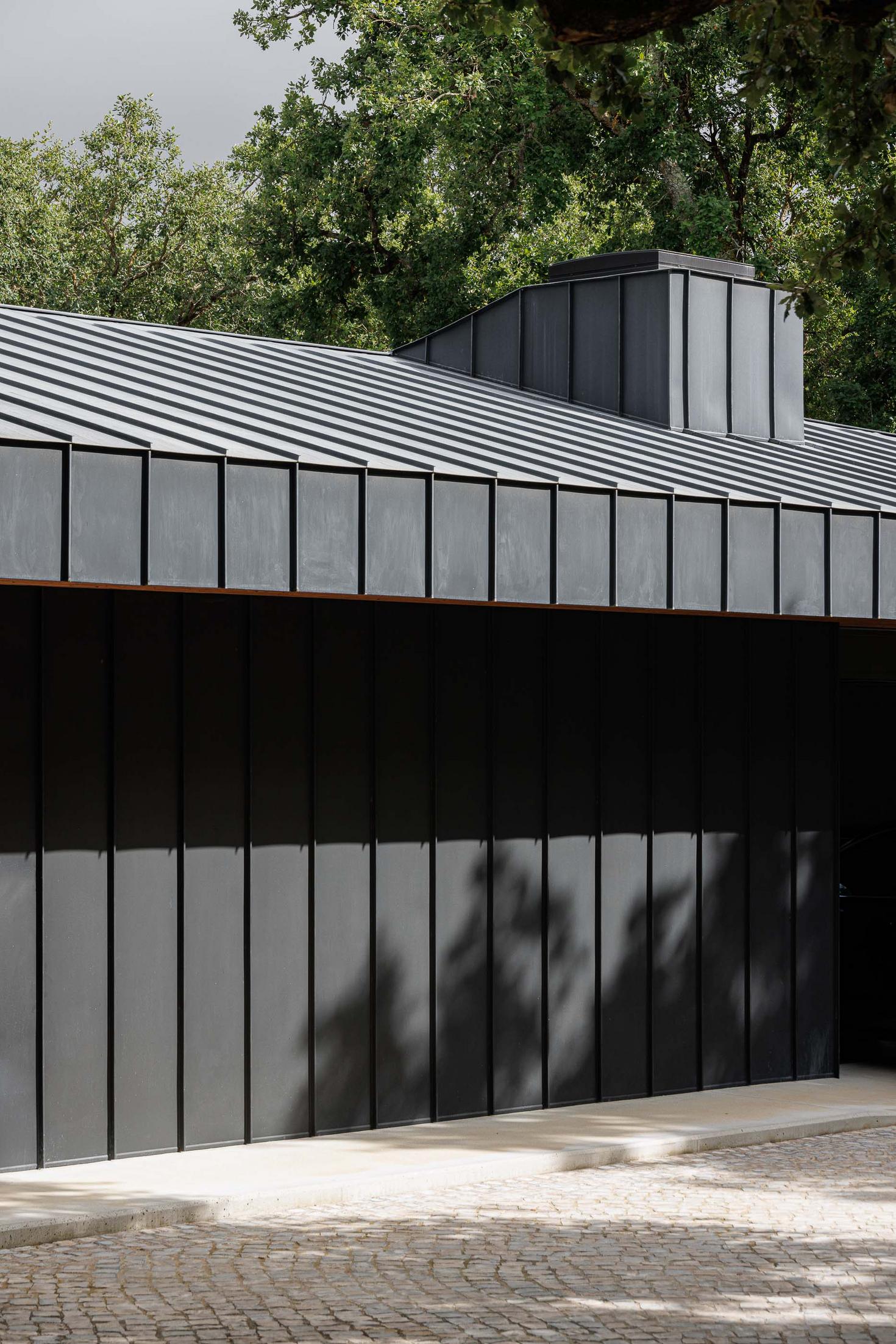 Fachada: Detalhe da fachada preta com revestimento metálico e telhado inclinado, pavimento em calçada portuguesa. — CC House – Quinta dos Carvalhos arquitetura Inception Architects Studio em Leiria