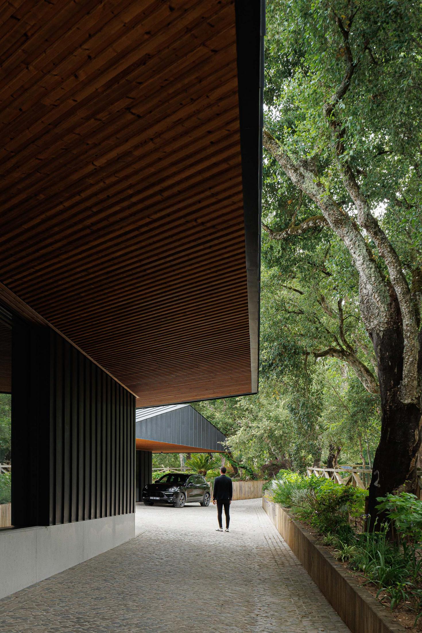 Exterior: Caminho de acesso à casa com revestimento em madeira no teto e fachada preta. — CC House – Quinta dos Carvalhos arquitetura Inception Architects Studio em Leiria