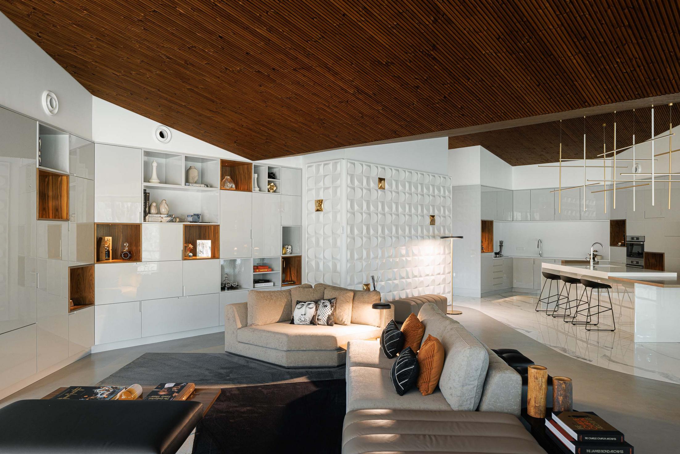 Sala de estar: Sofás bege, estantes brancas com detalhes em madeira, parede com revestimento branco texturado e cozinha ao fundo. — CC House – Quinta dos Carvalhos arquitetura Inception Architects Studio em Leiria