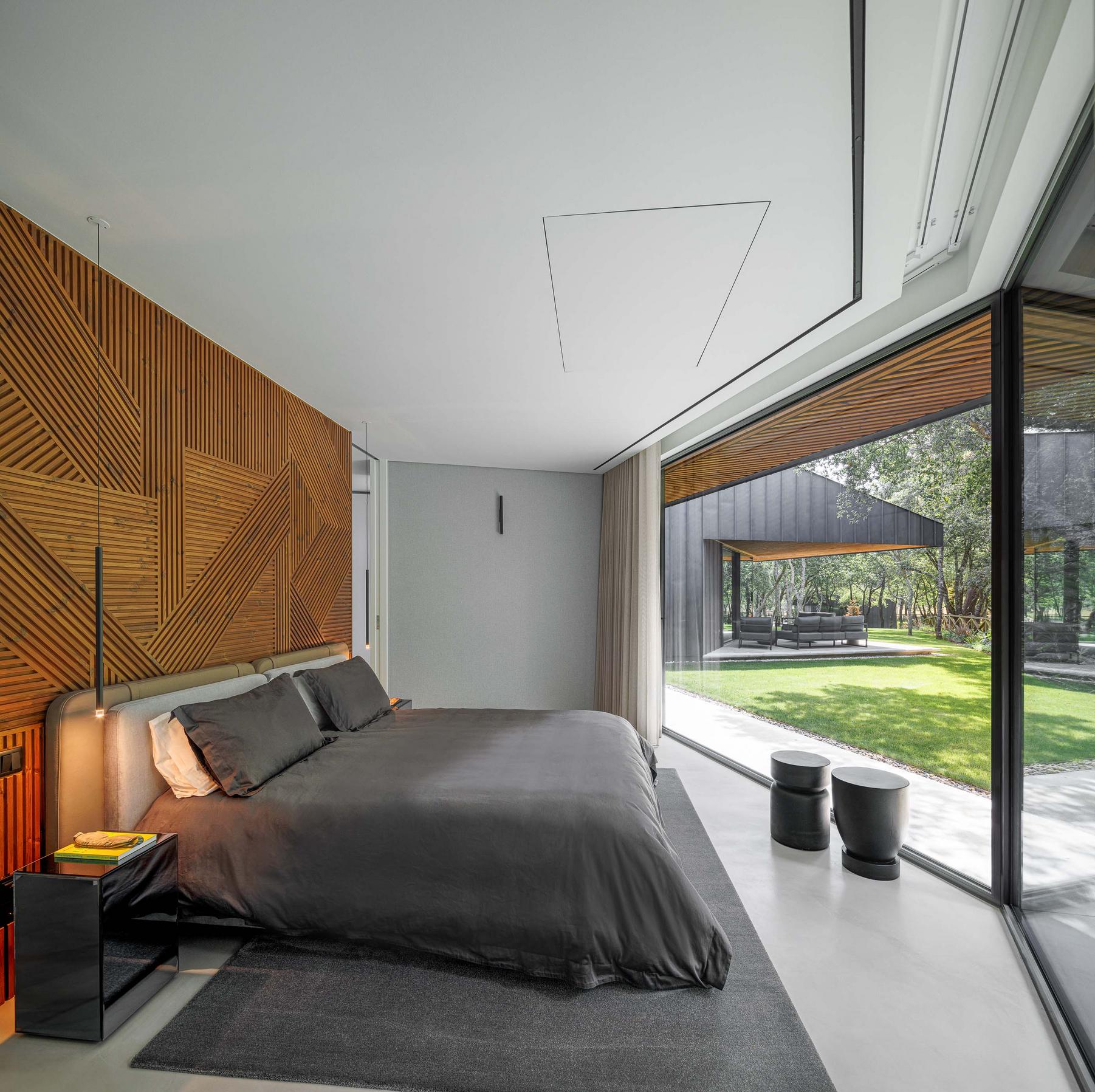 Quarto: Cama com roupa cinzenta, parede em madeira com padrão geométrico e vista para o jardim. — CC House – Quinta dos Carvalhos arquitetura Inception Architects Studio em Leiria