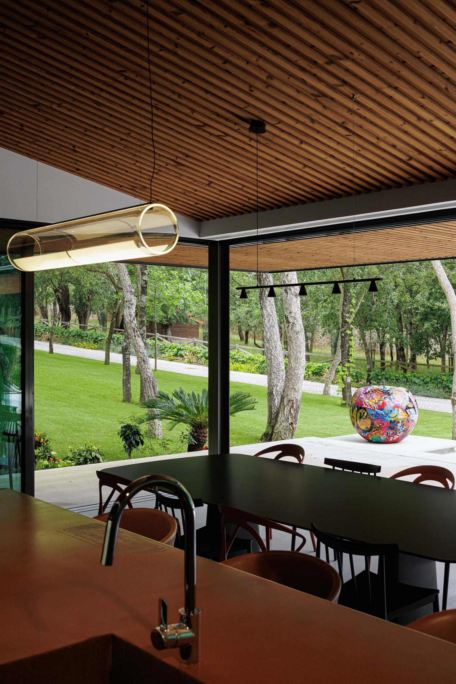 Cozinha/Sala de jantar: Vista da cozinha para o exterior, mesa de jantar com cadeiras e candeeiro tubular suspenso. — CC House – Quinta dos Carvalhos arquitetura Inception Architects Studio em Leiria