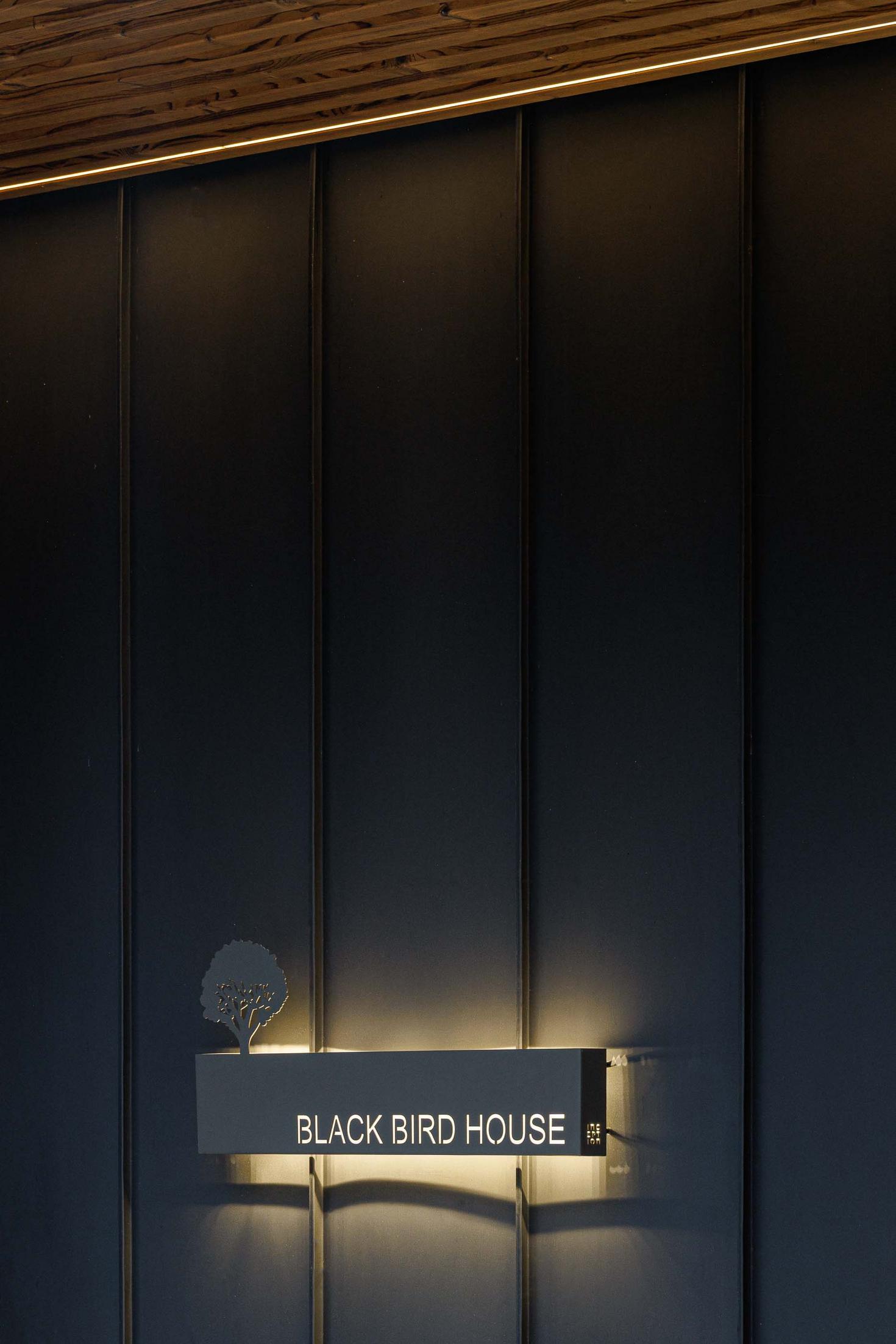 Entrada: Placa com o nome "Black Bird House" iluminada, parede com revestimento vertical preto e teto em madeira. — CC House – Quinta dos Carvalhos arquitetura Inception Architects Studio em Leiria