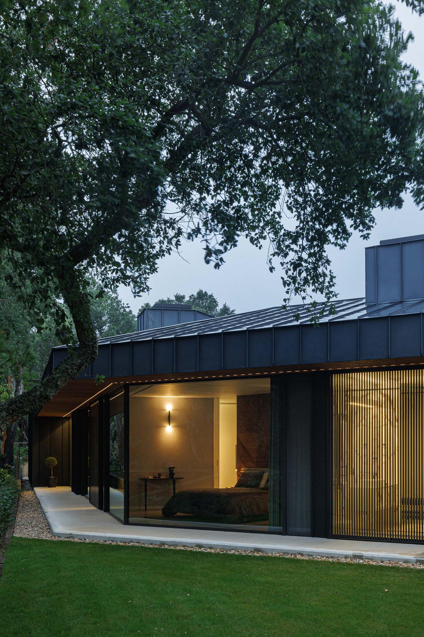 Exterior: Vista da casa a partir do jardim, com quarto visível através da janela e revestimento em madeira. — CC House – Quinta dos Carvalhos arquitetura Inception Architects Studio em Leiria