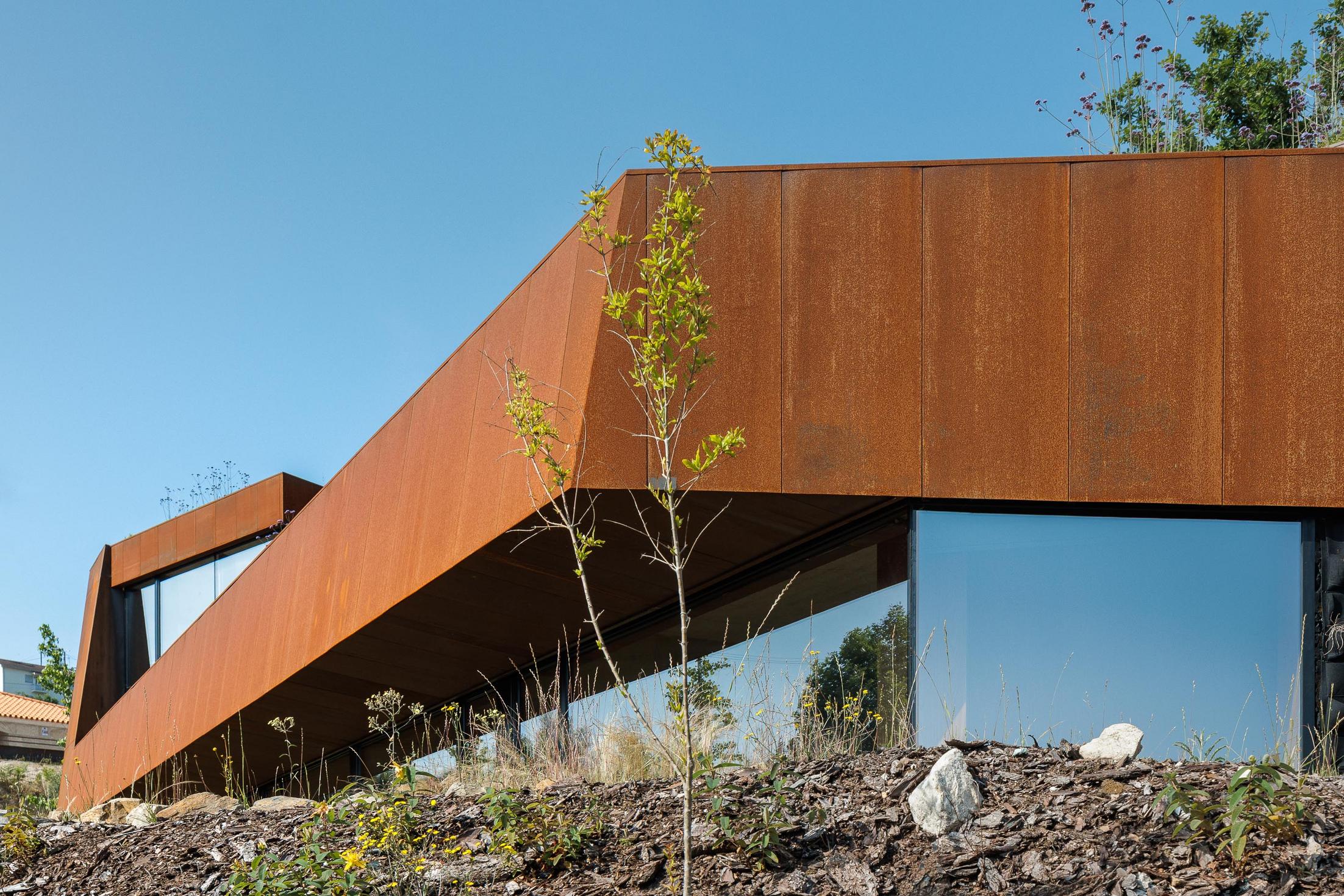 Exterior: Detalhe da fachada em aço corten, com vegetação rasteira e uma pequena árvore em primeiro plano. — Casa Corten arquitetura HPA Arquitetura e Investimentos em Celorico de Basto